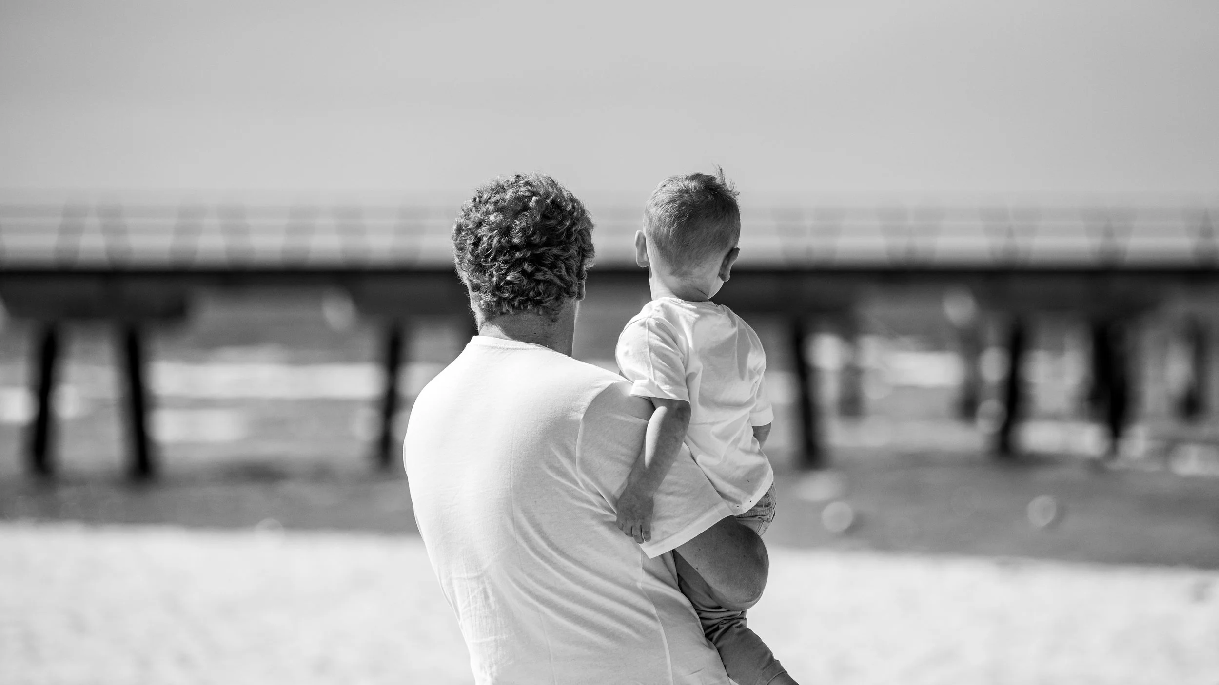 A man with curly hair holding a young boy on a beach, facing a pier in the background in black and white.
