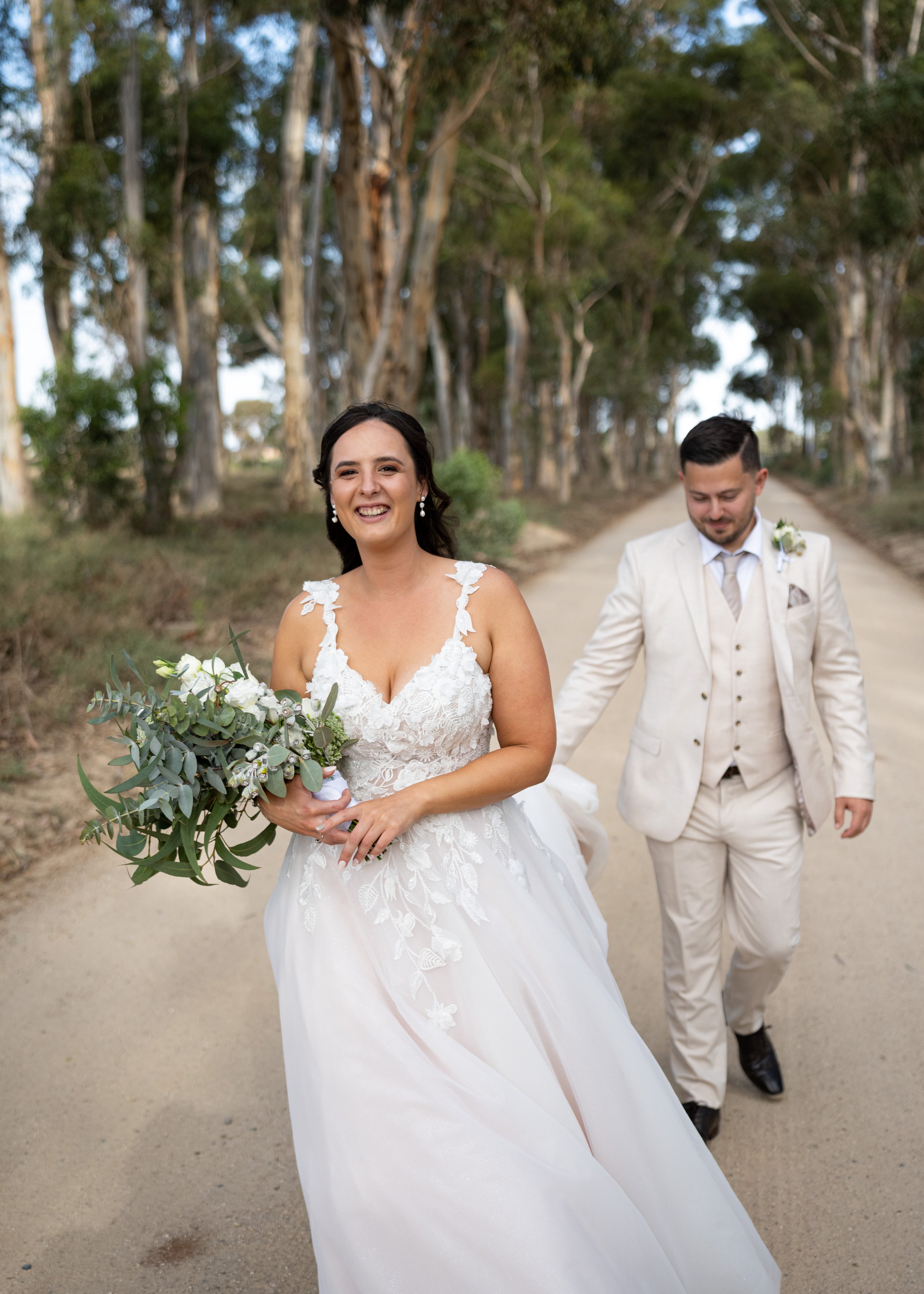 A bride in a white wedding dress holding a bouquet of greenery and white flowers, smiling, walking with a groom in a light-colored suit on a dirt road surrounded by trees.