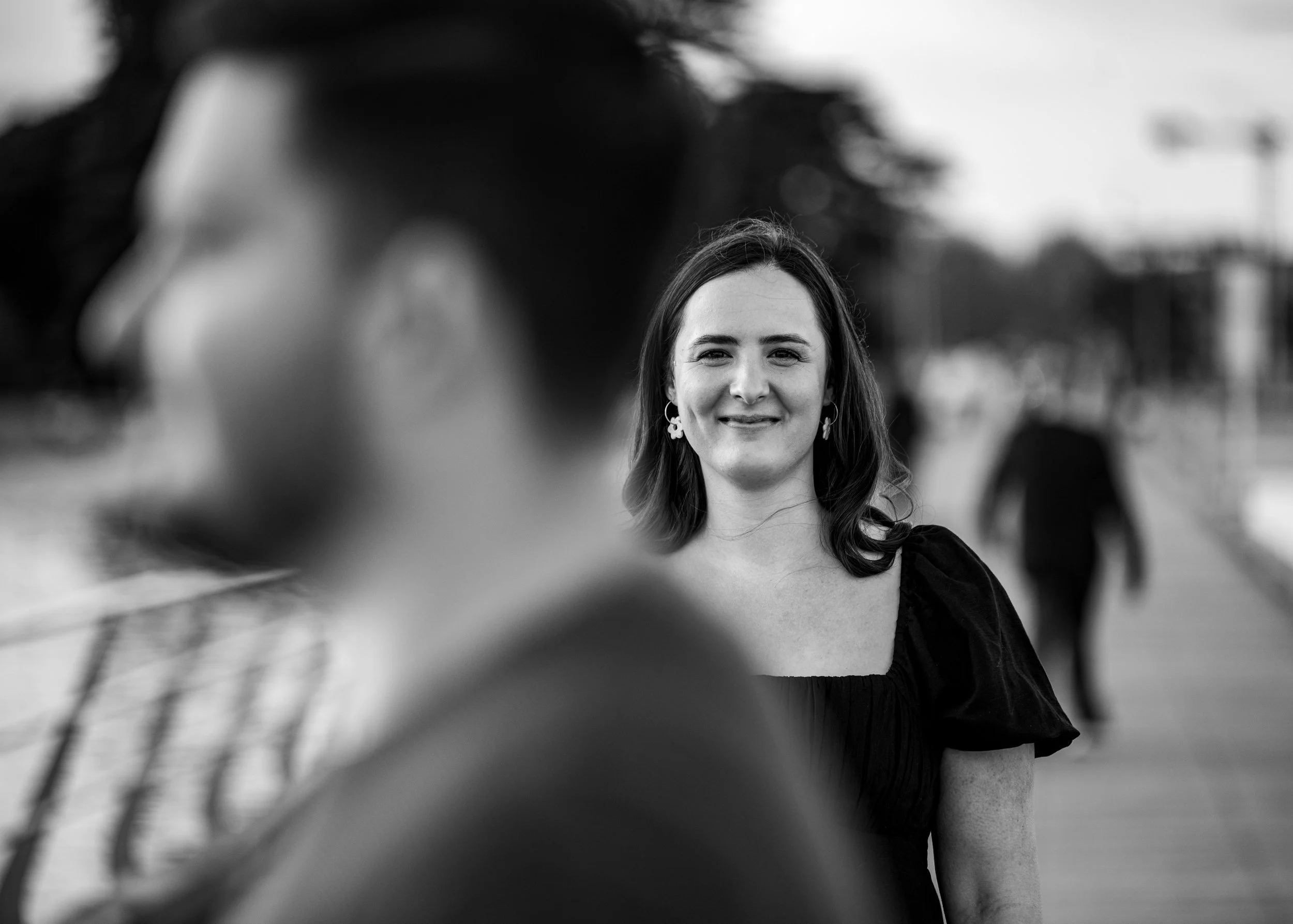 A woman in focus smiling at the camera, with another person blurred in the foreground. The scene is outdoors near a walkway, possibly by a river or waterfront, with trees and structures in the background.