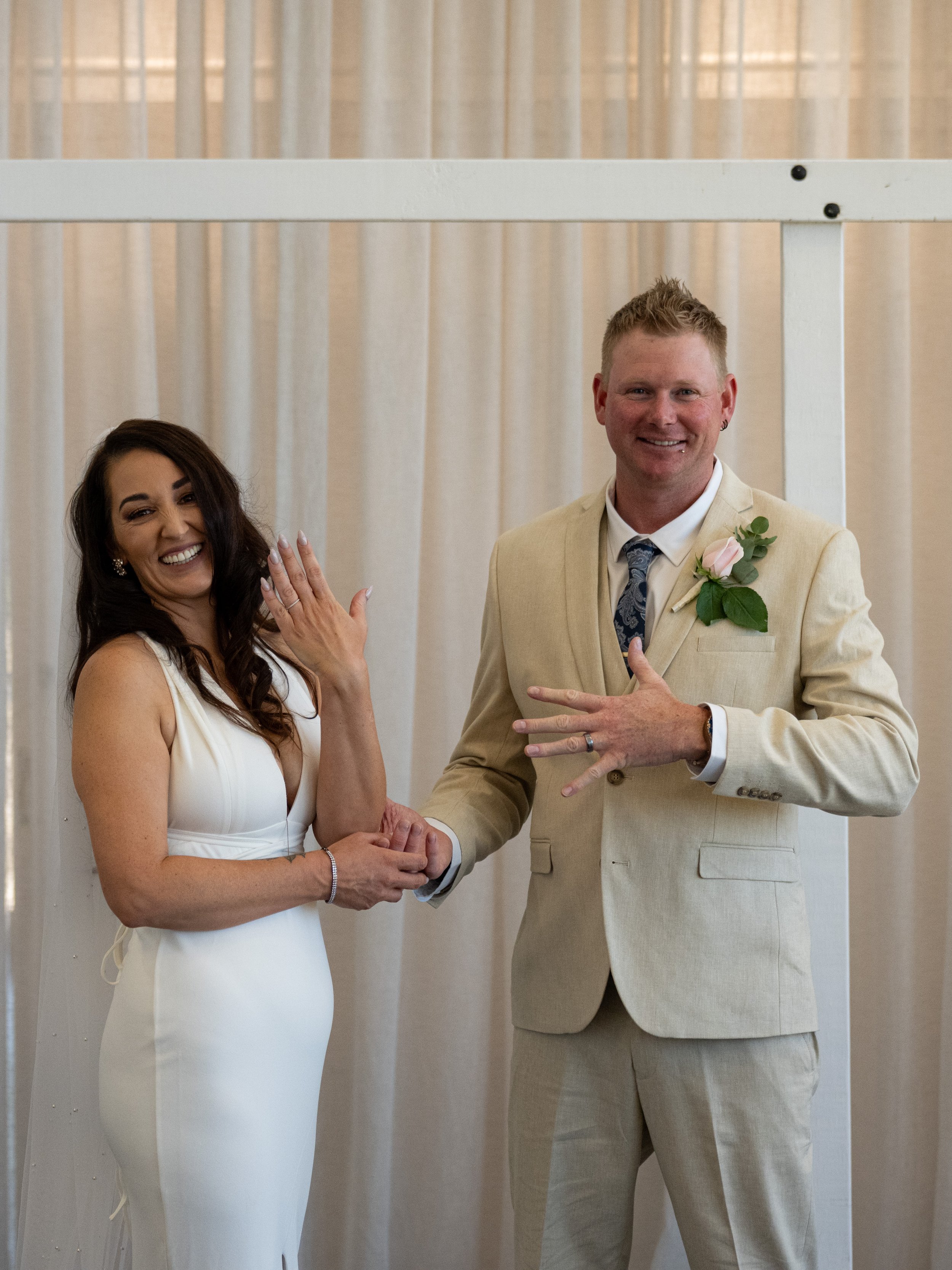 A couple standing at their wedding, holding hands, with the bride showing her engagement ring and the groom displaying his wedding band, smiling in front of cream-colored curtains.