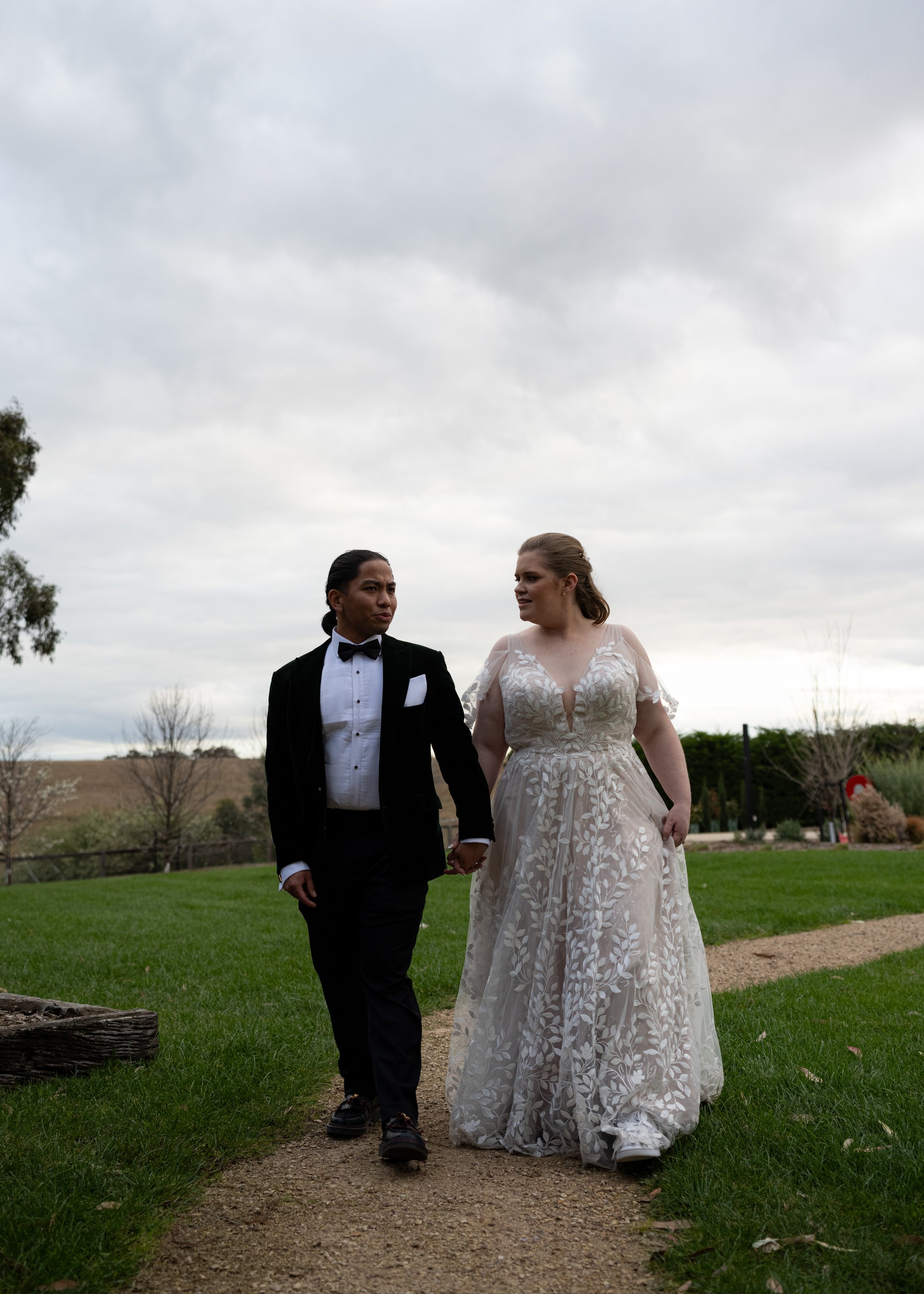 A couple dressed in formal wedding attire walking hand in hand outdoors under a cloudy sky.
