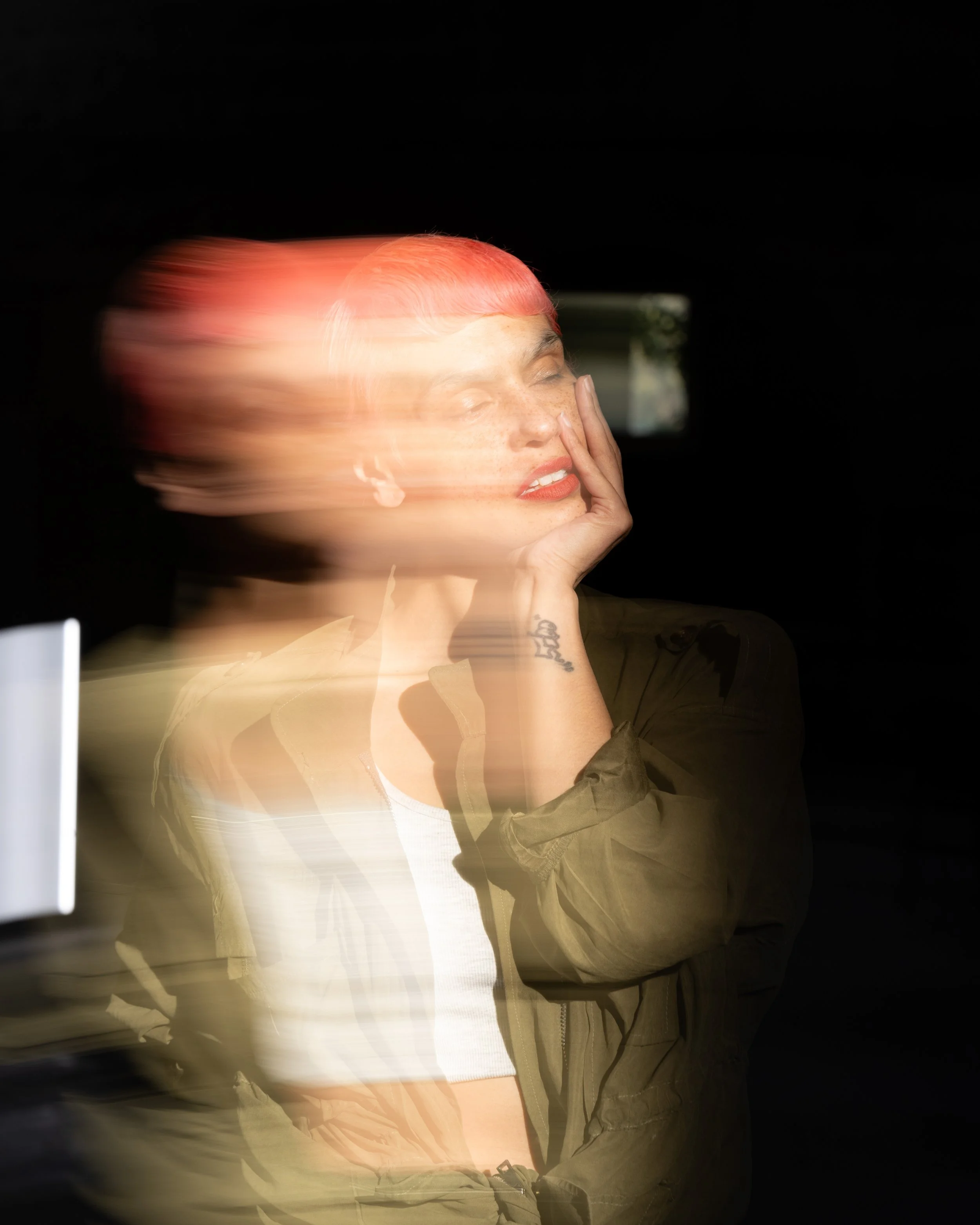 A woman with pink hair and a tattoo on her wrist has her hand on her face with her eyes closed. There is motion blur around her face against a dark background.