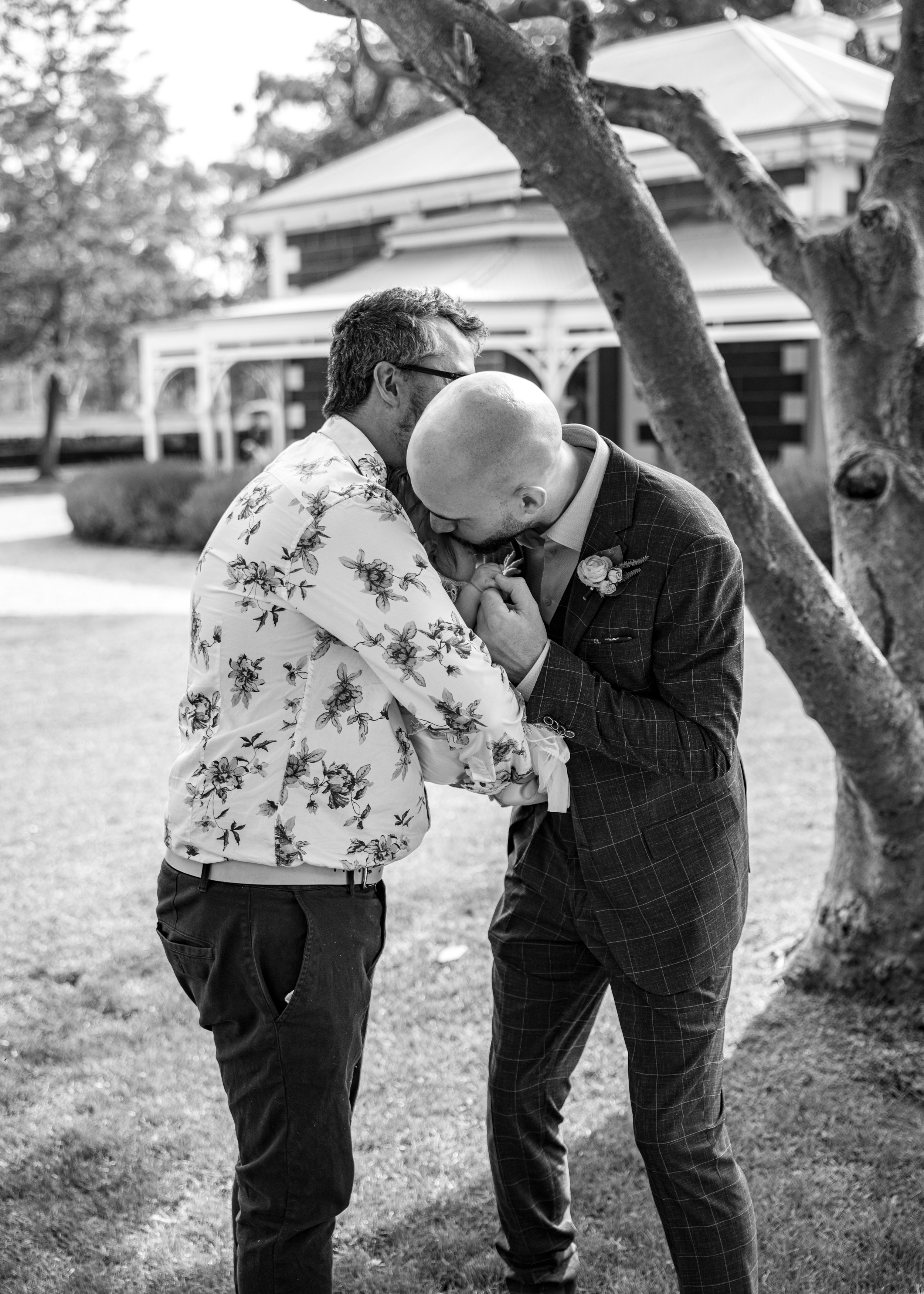 Two men dressed in formal attire gently holding and kissing a baby under a tree, outdoors, during a wedding or special event.
