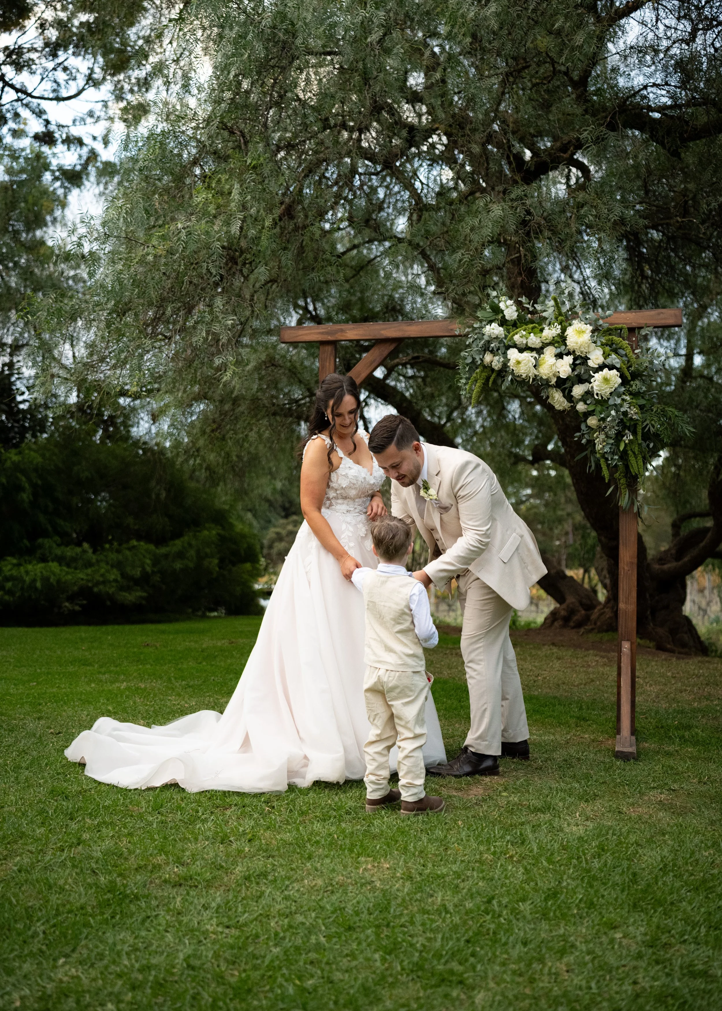 A wedding ceremony outdoors with a bride, groom, and a young boy under a wooden arch decorated with white flowers and greenery, set on a grassy area with trees in the background.