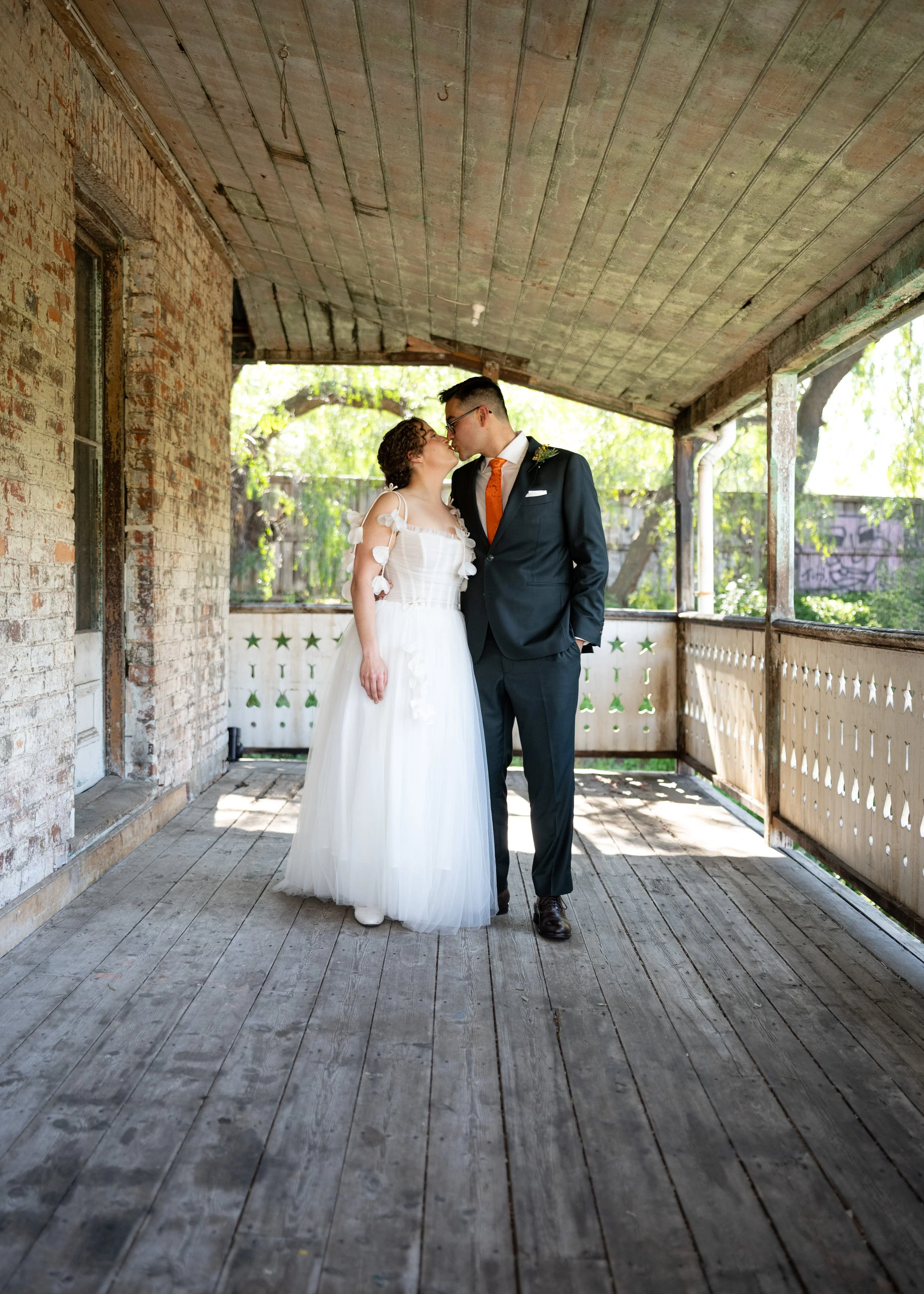 A bride and groom standing on a worn wooden porch, sharing a close moment and leaning in for a kiss, surrounded by greenery and natural light.