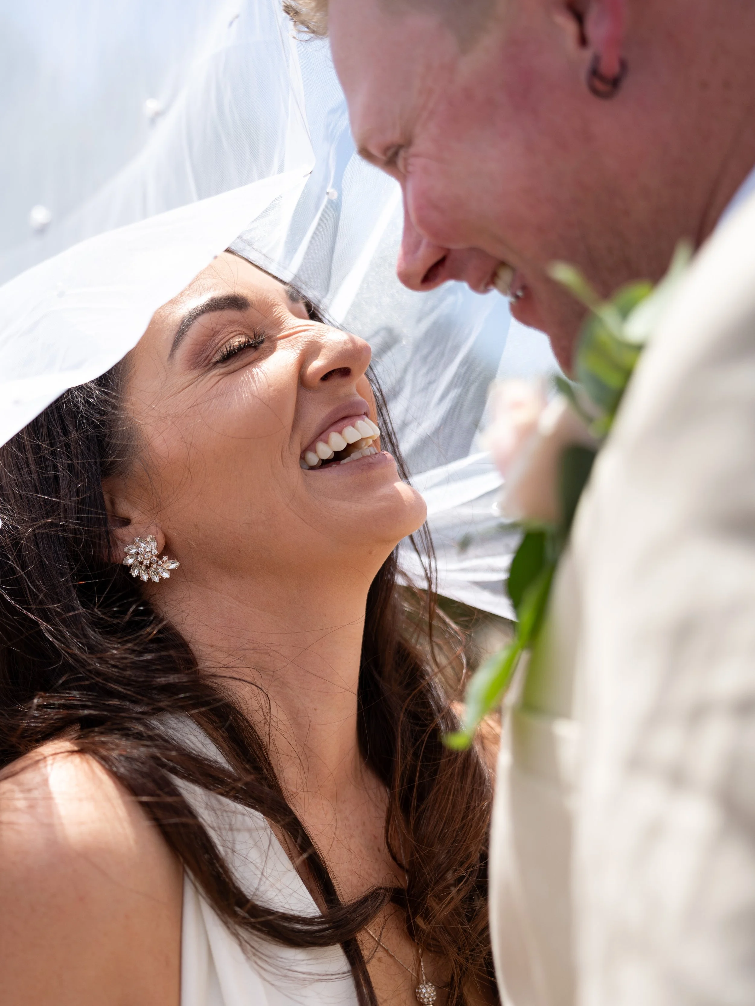 A bride and groom sharing a joyful moment under a clear umbrella, smiling at each other during their wedding celebration.