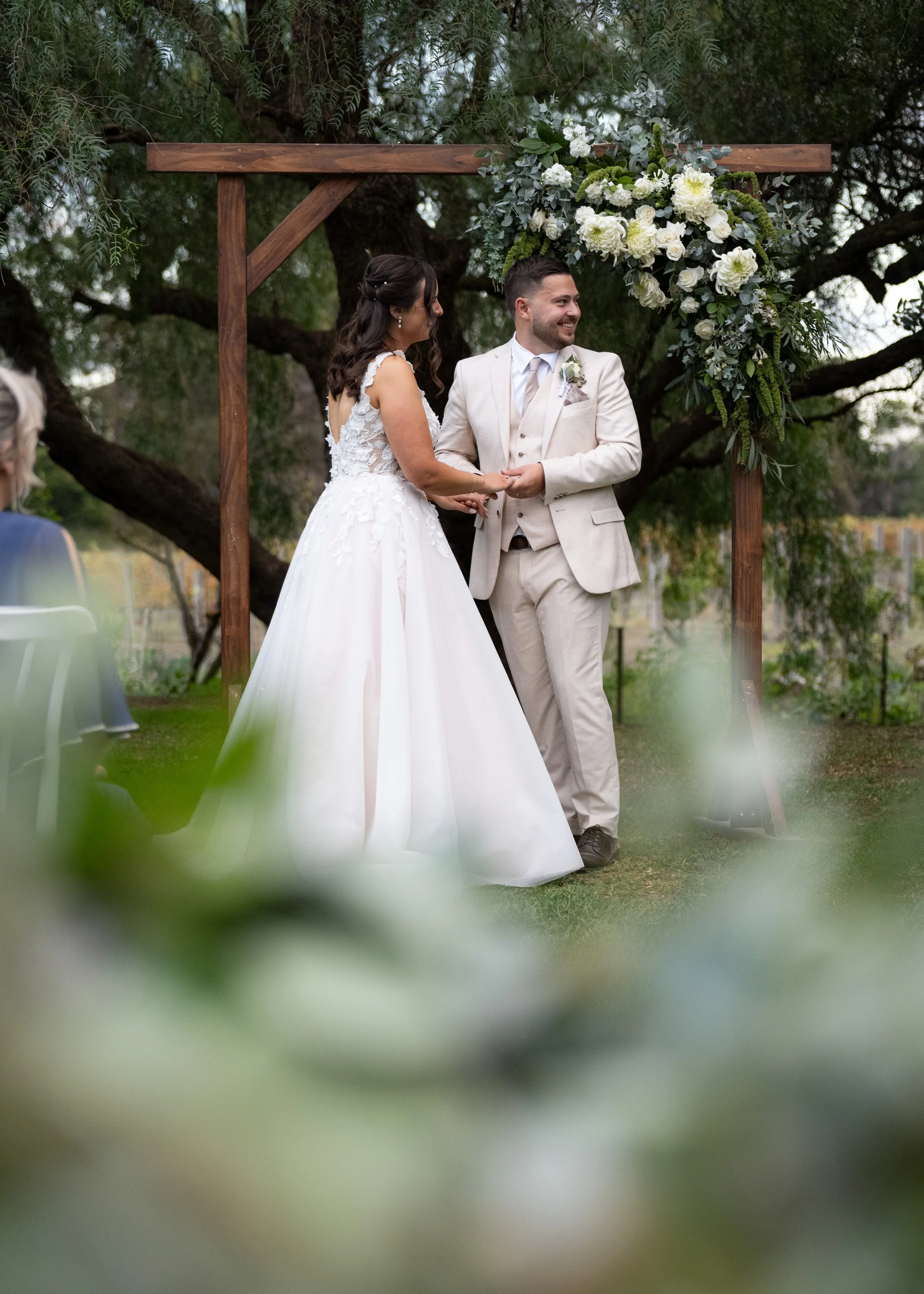 A bride and groom holding hands during their outdoor wedding ceremony under a wooden arch decorated with white and green flowers, with trees and greenery in the background.