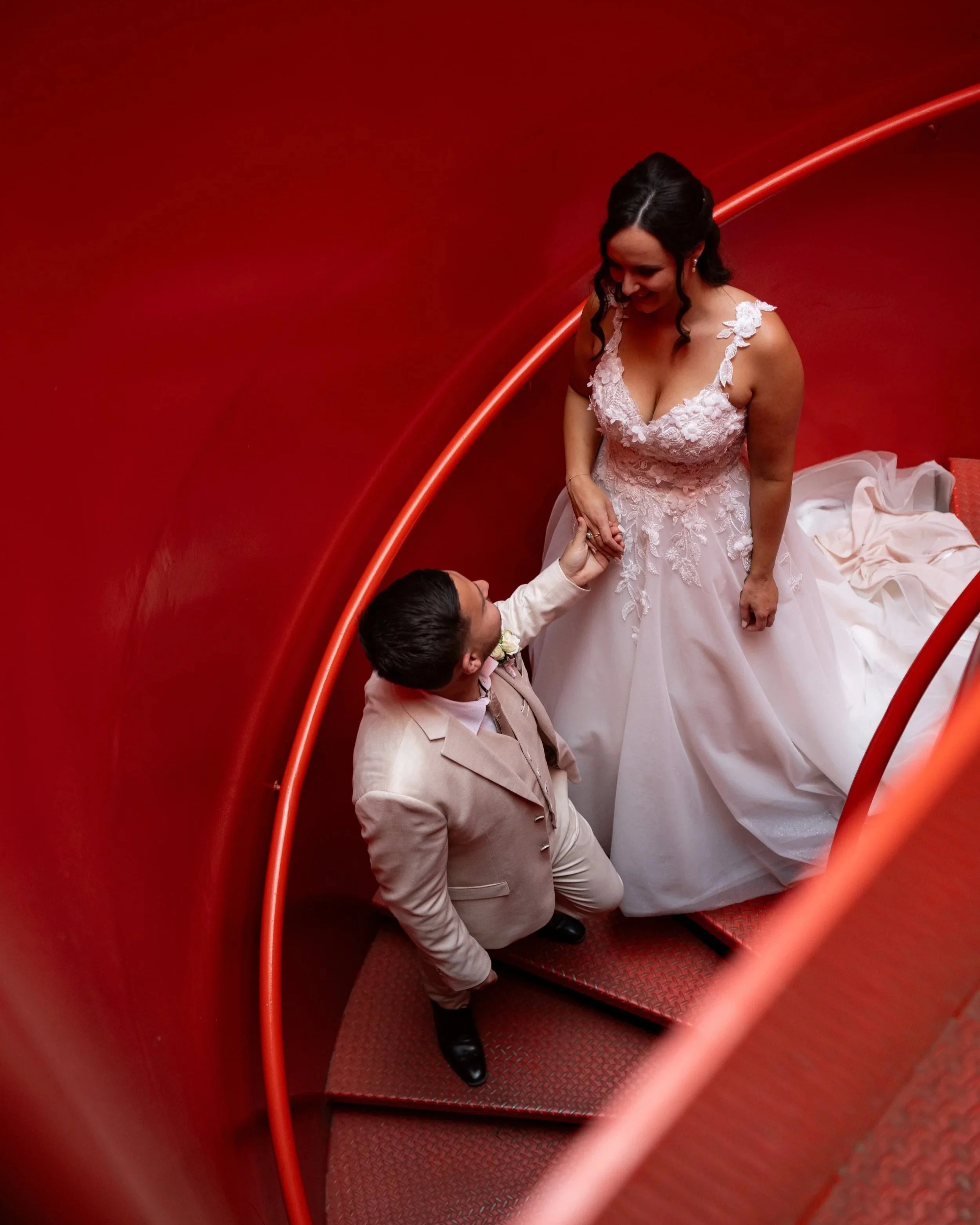 A bride and groom stand on a red spiral staircase, holding hands and looking at each other happily a Shadowfax Winery wedding in Melbourne