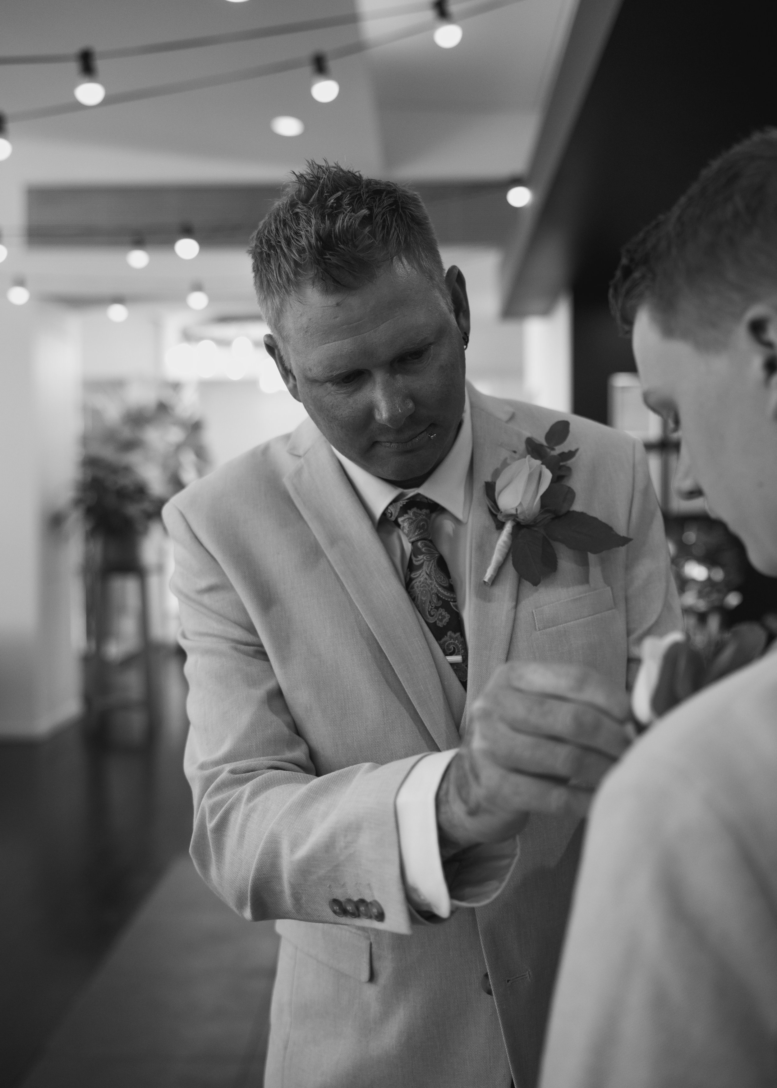 A man in a light-colored suit pinning a boutonnière on another man at a wedding.