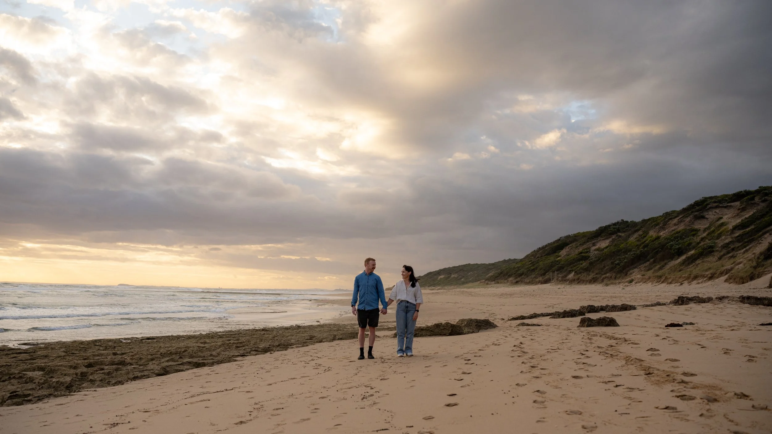 A couple walking along the beach at sunset, holding hands with a cloudy sky and sand dunes in the background.