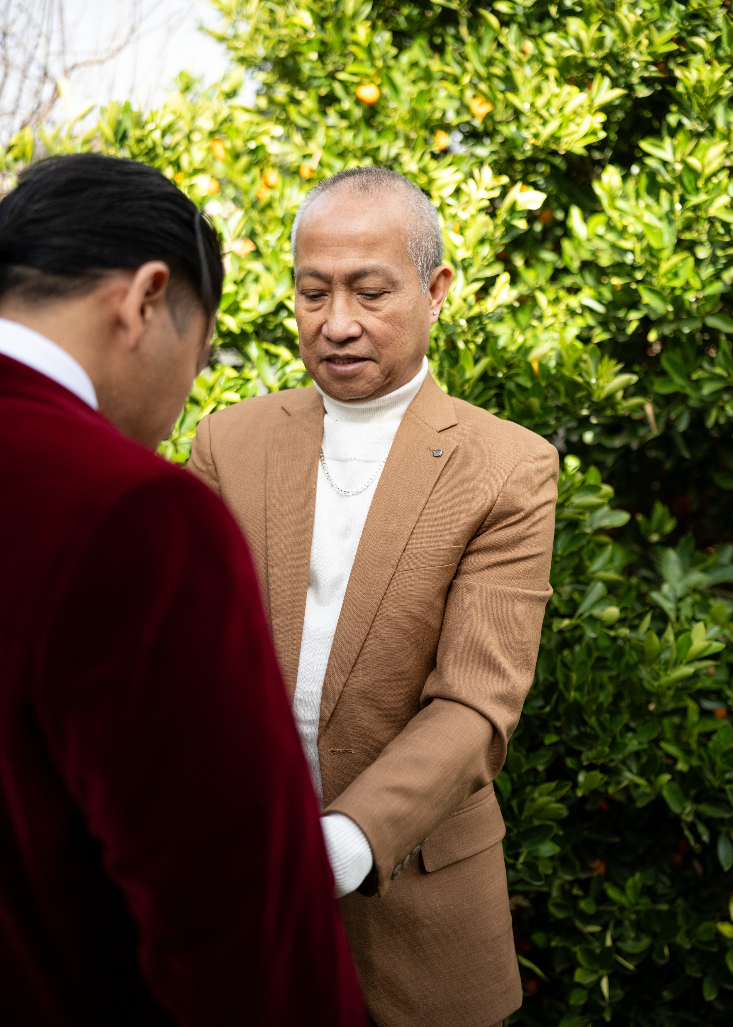 Two men standing outdoors, one with a shaved head wearing a tan suit and white turtleneck, and the other with dark hair in a red velvet jacket, engaging in a conversation near green bushes with orange flowers.
