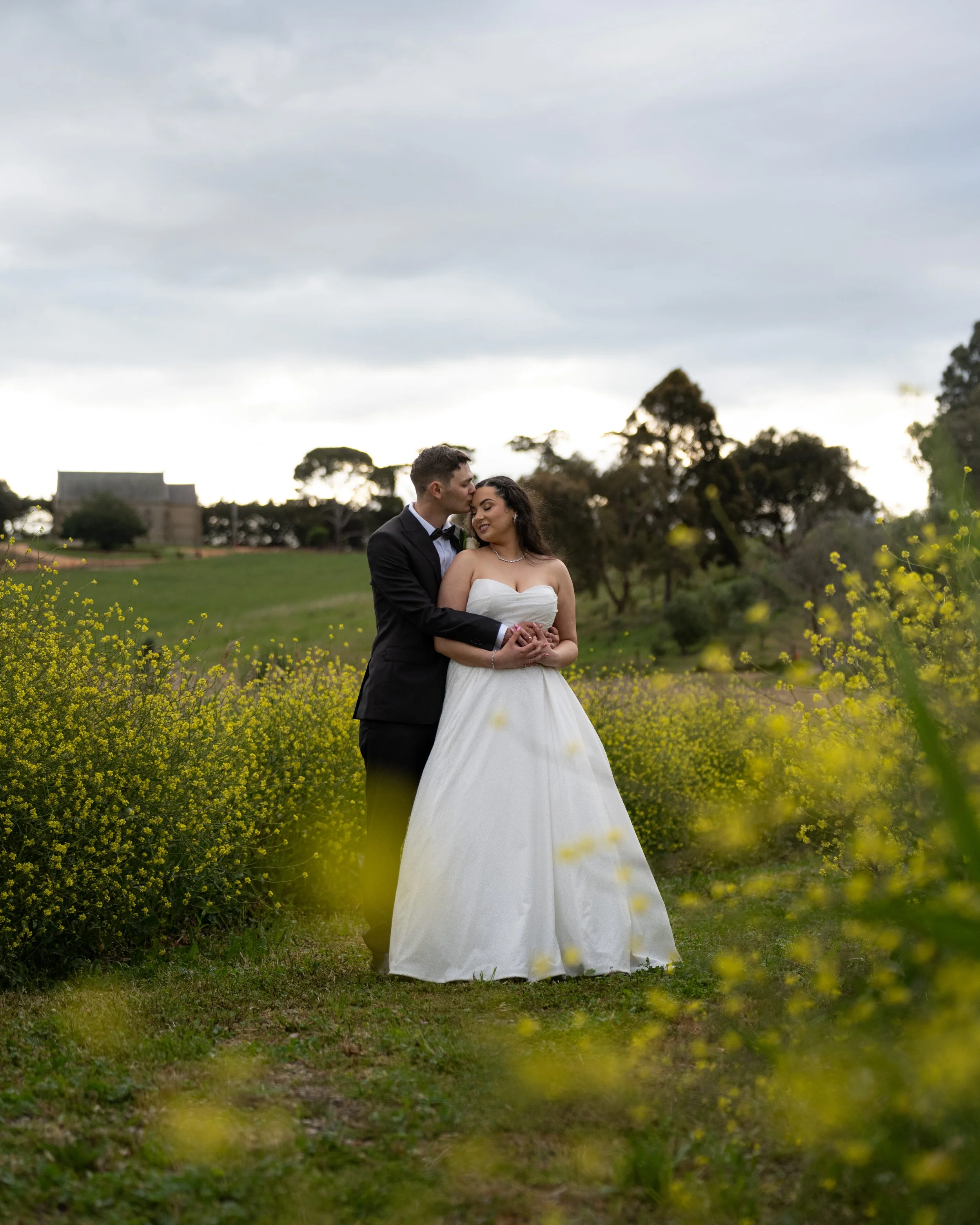 A newlywed couple stands in a field of yellow flowers during the daytime, with trees at Barrabool Maze Estate Geelong.