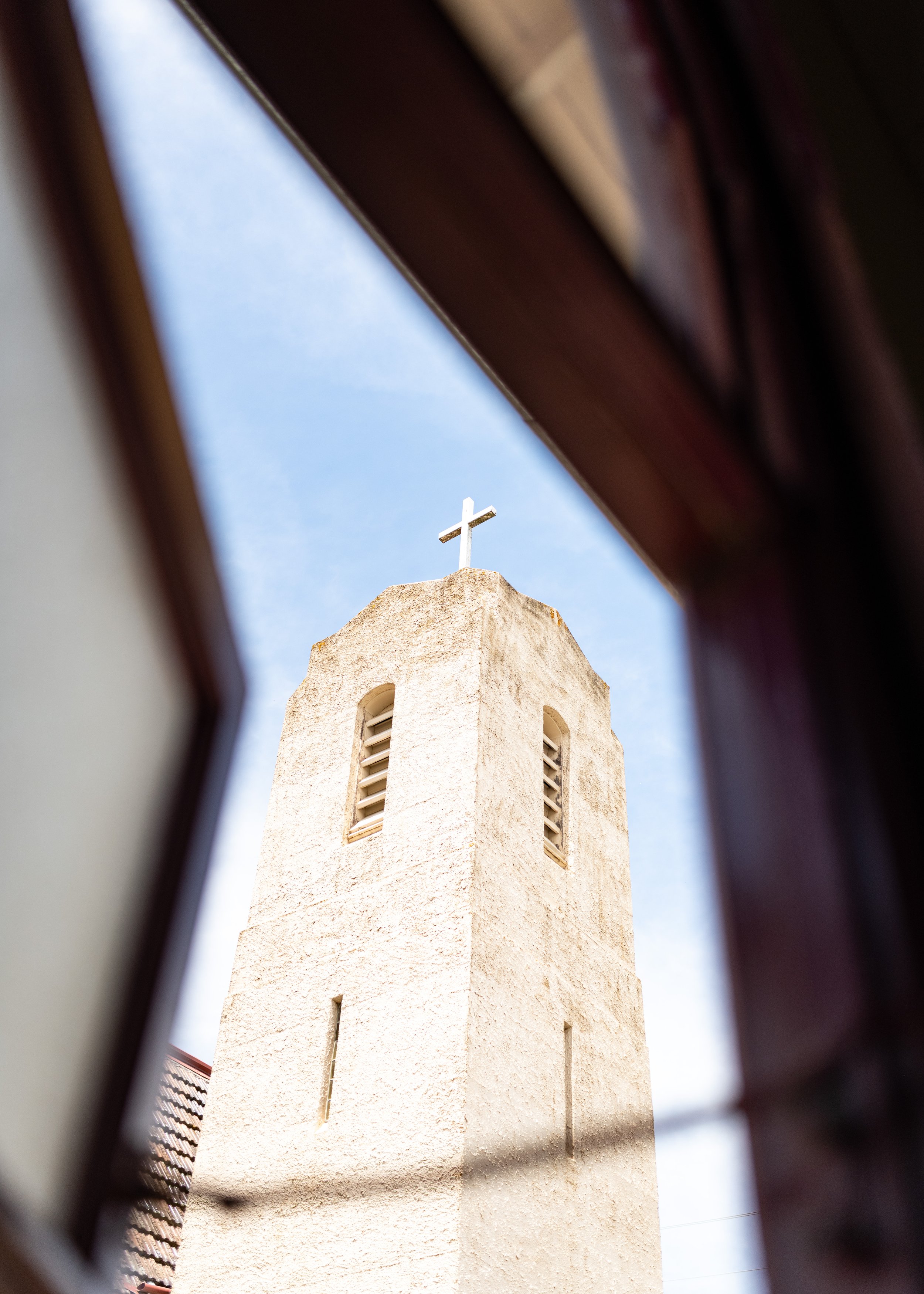 View of a church steeple with a cross on top, seen through a window frame, against a blue sky.