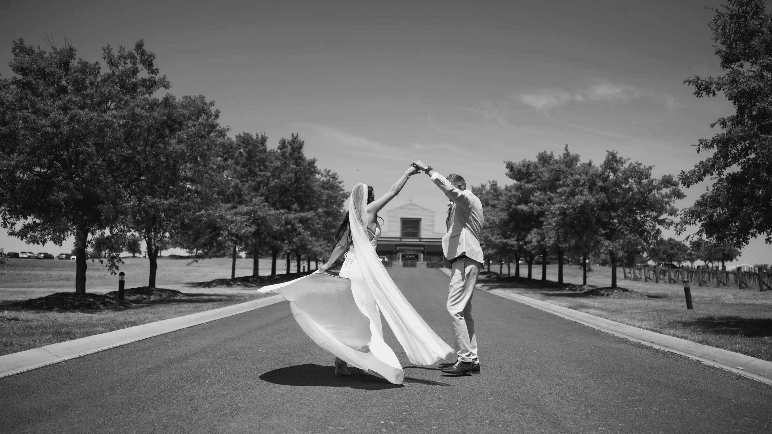 A bride and groom dancing on an empty driveway surrounded by trees, with a building in the background, captured in black and white.