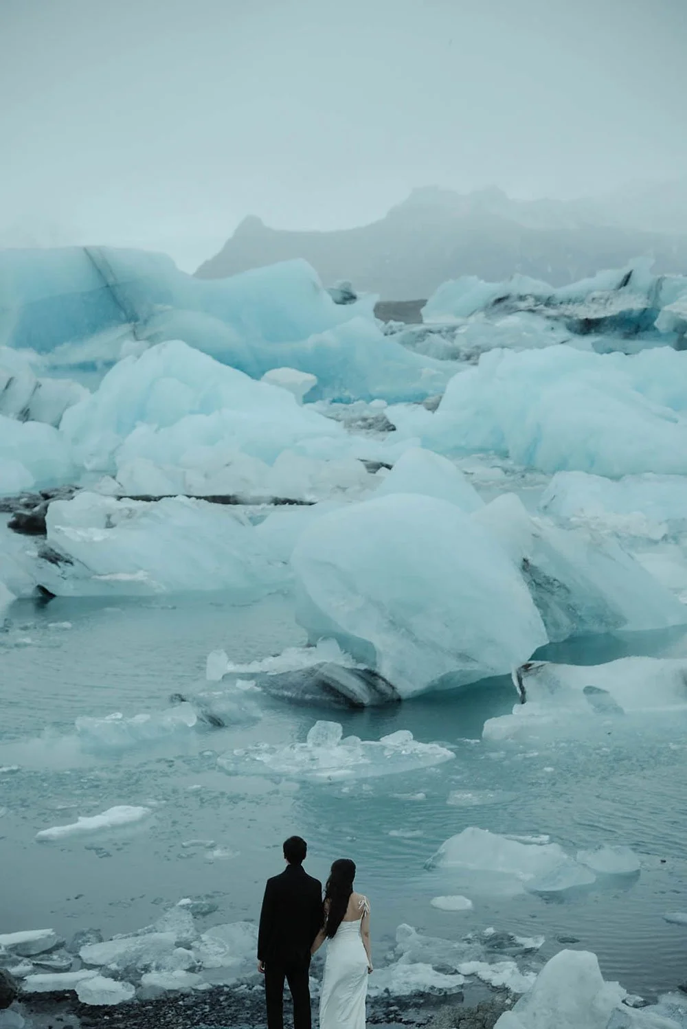 A couple stands beside floating blue ice at an Iceland glacier lagoon during their adventurous elopement.
