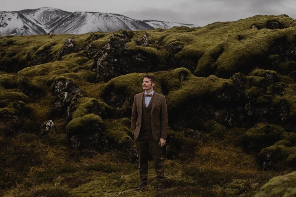 A groom stands among moss-covered lava rocks with snow-dusted Icelandic mountains behind him.