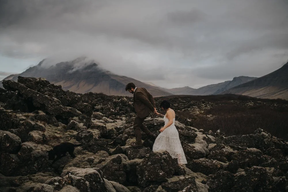 An eloping couple walks together through a rugged volcanic landscape beneath Icelandic mountains.
