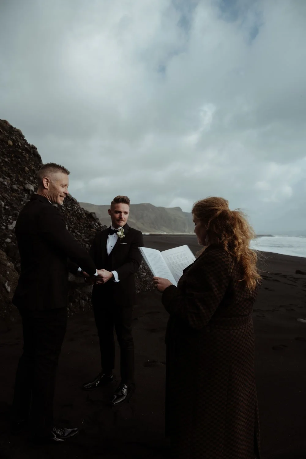 Two grooms hold hands during their Iceland elopement ceremony on a windswept black sand beach.