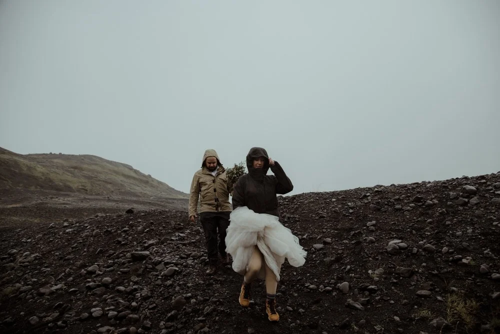 A bride lifts her dress while hiking through wet volcanic terrain during an adventurous Iceland elopement.