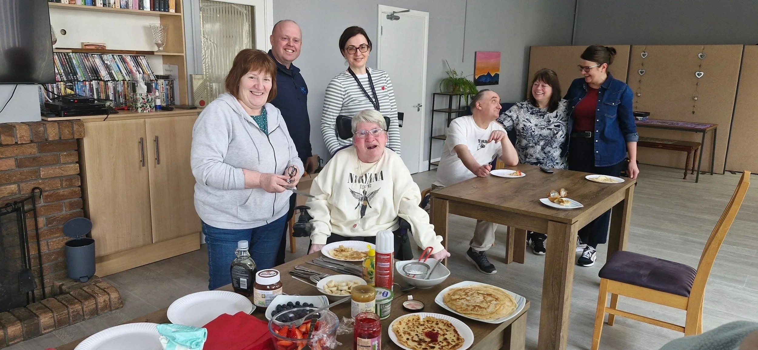 A group of eight people, mostly women, gathered around tables with food in a room with a brick fireplace. They are smiling and appear to be celebrating or enjoying a meal together.