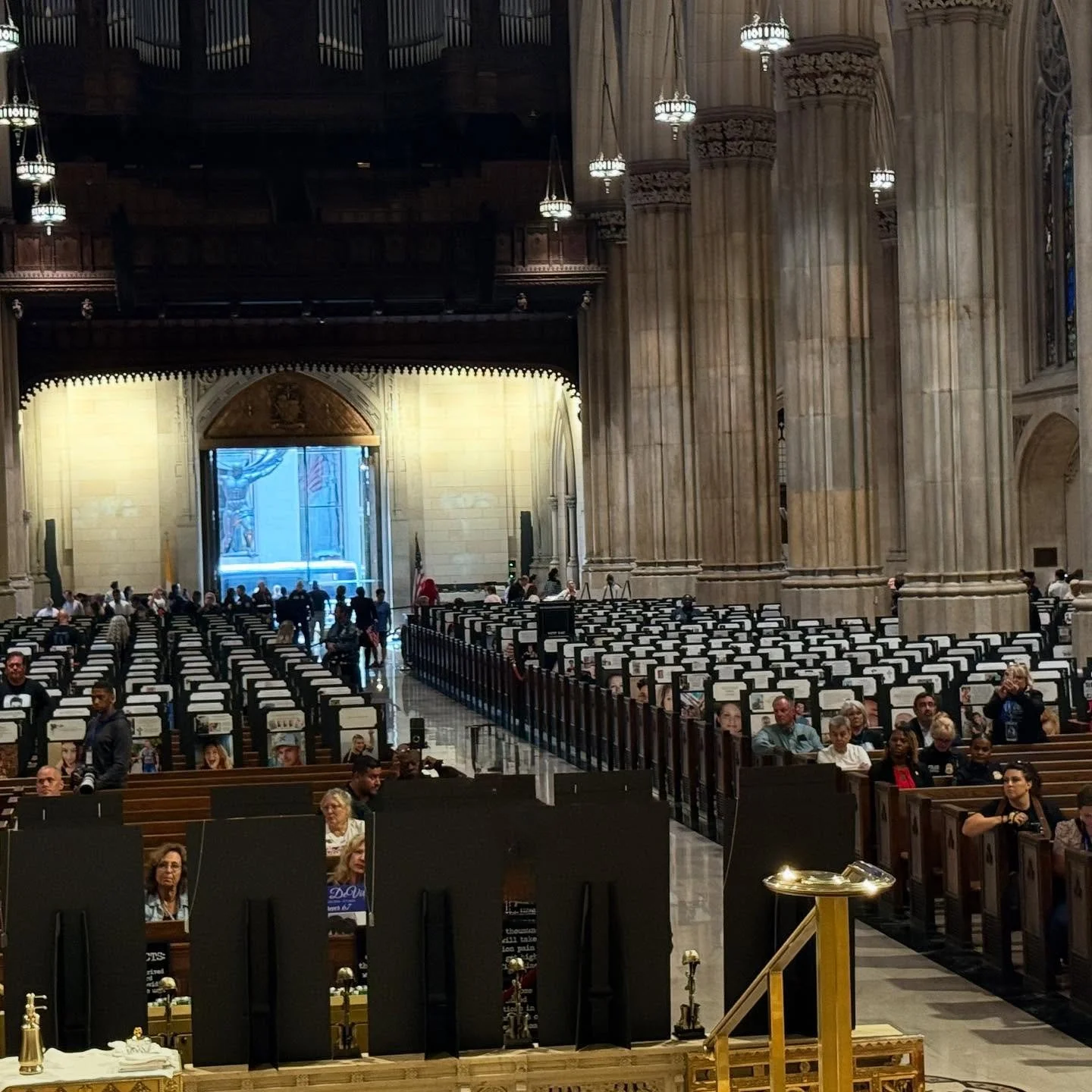 Interior of a large church or cathedral with rows of chairs and people seated, high stone columns, an altar at the front, stained glass windows, and ornate lighting fixtures hanging from the ceiling.