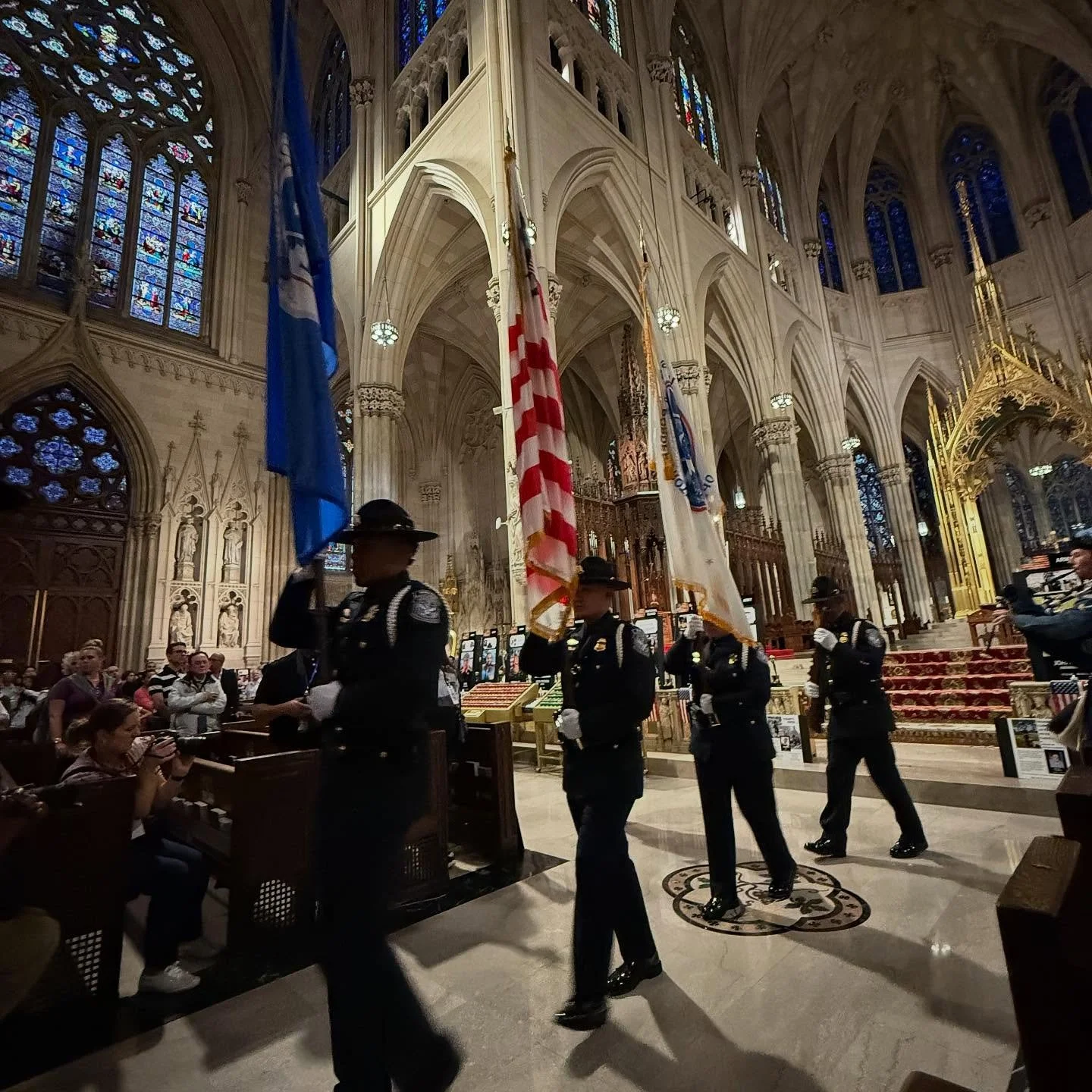 Uniformed police officers carrying flags walk through the interior of a grand, ornate church or cathedral with stained glass windows and high vaulted ceilings, while a seated audience observes the procession.