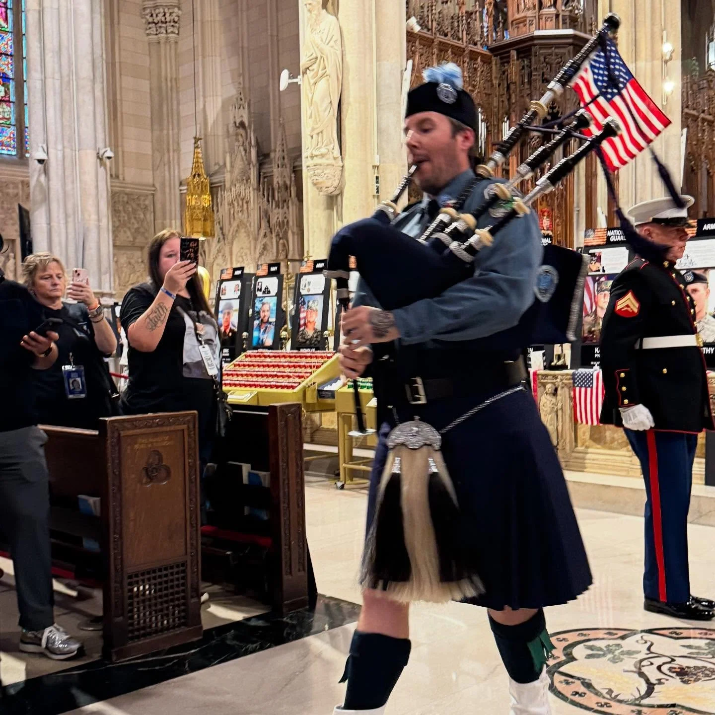 A man dressed in traditional Scottish attire playing bagpipes inside a grand church or cathedral with people observing, including women recording and taking photos, and a Marine standing at attention in dress uniform.