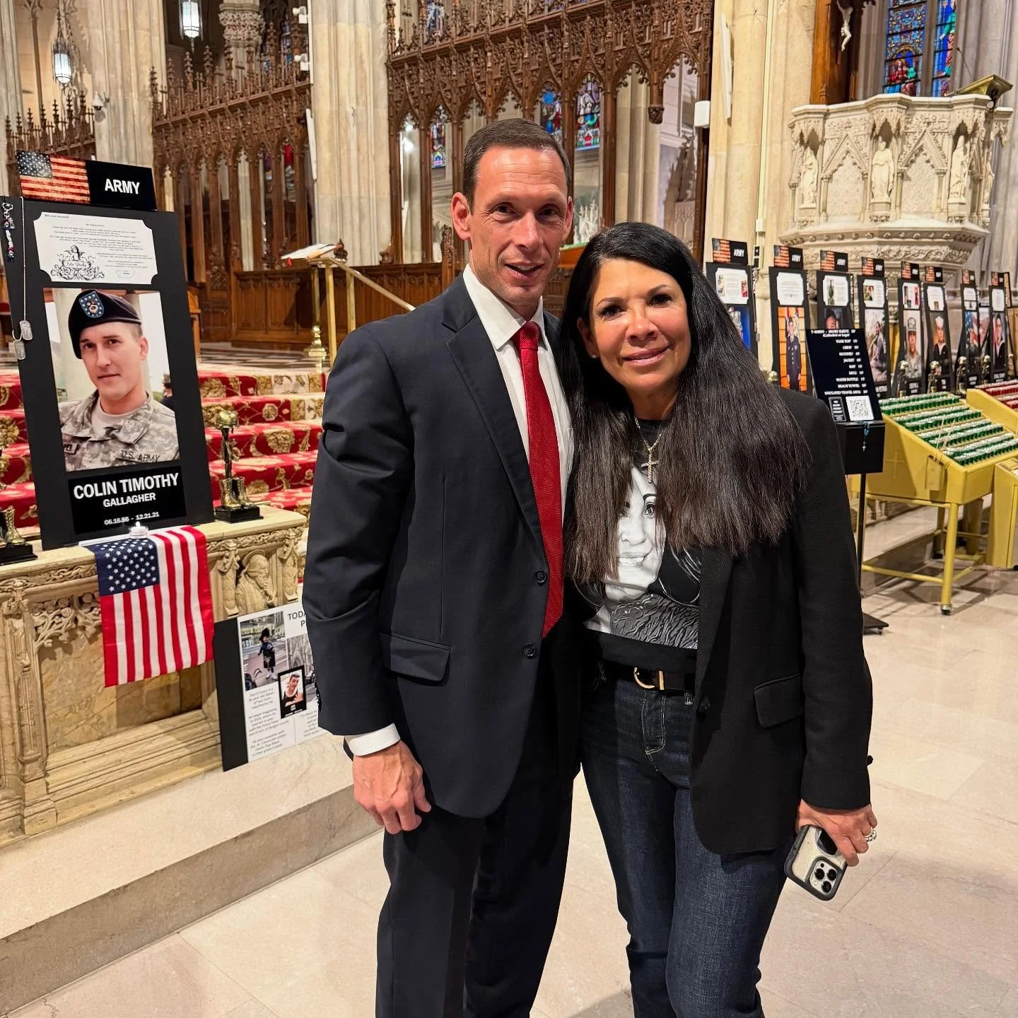 A man in a dark suit, white shirt, and red tie standing with a woman in a black blazer and jeans inside a church or cathedral. The background includes memorial displays with photos of soldiers, a small American flag, and religious stained glass windo