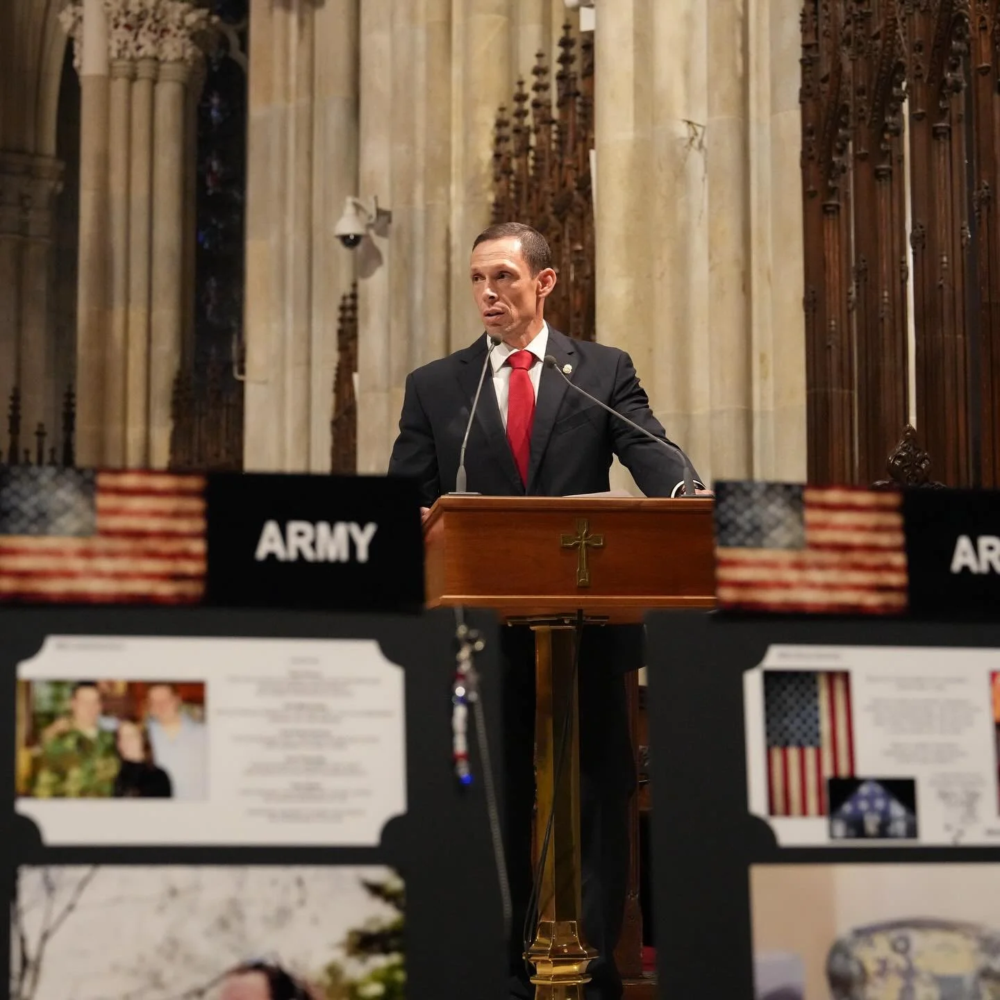 A man in a dark suit and red tie speaking at a wooden podium inside a church, with flags and informational posters about the Army on the table in front.
