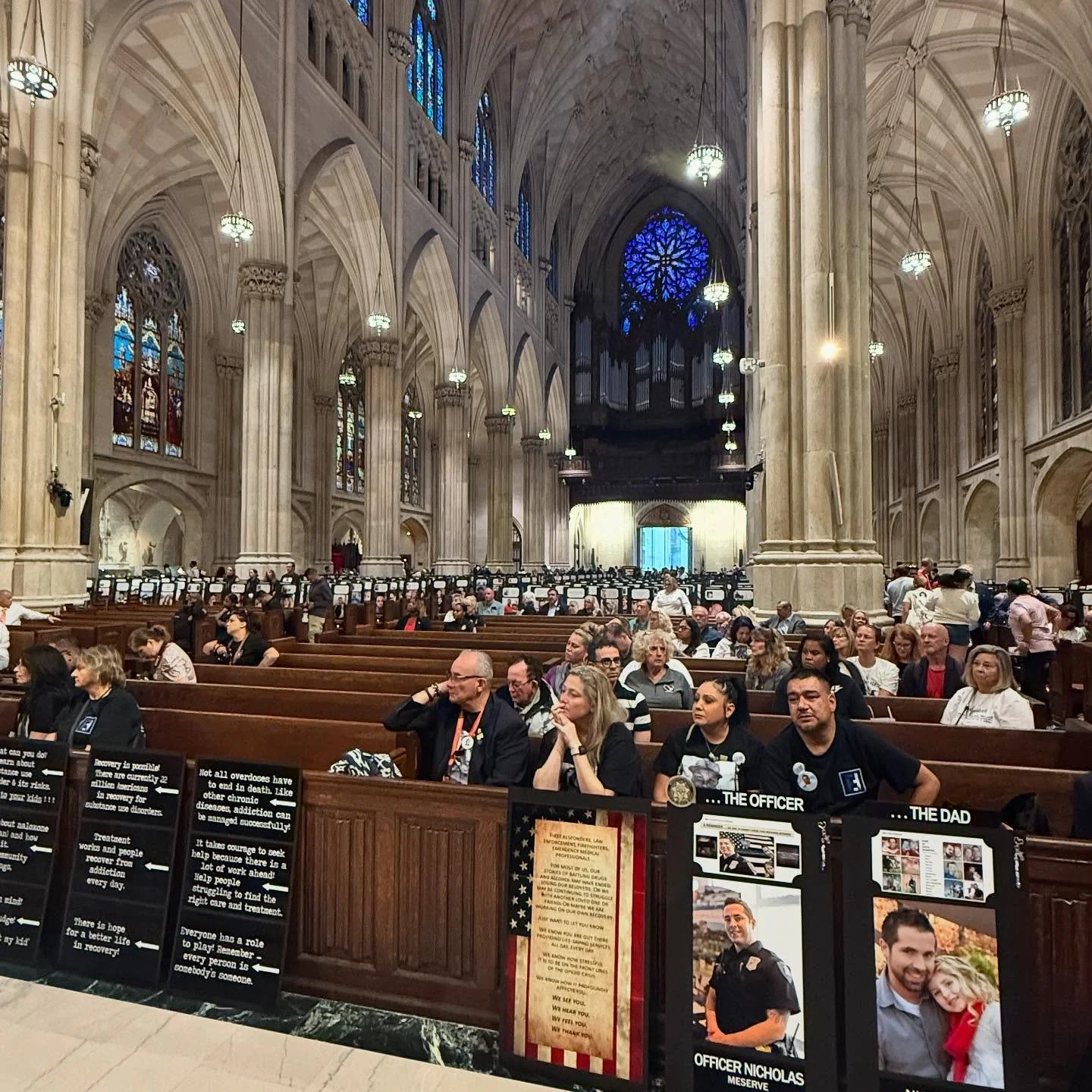 Inside a cathedral with high vaulted ceilings, stained glass windows, and hanging lights. People are seated in pews and attending a ceremony or event.