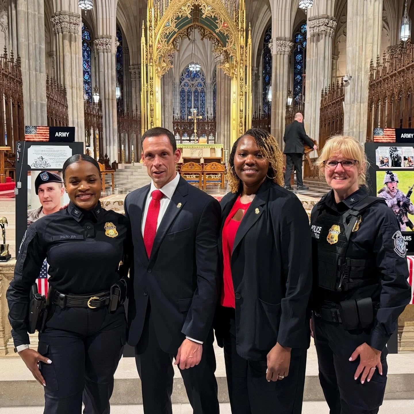 Four people standing inside a grand cathedral, with two women in police uniforms on either side and a man and woman in formal attire in the middle. The background features ornate architecture, stained glass windows, and an altar.