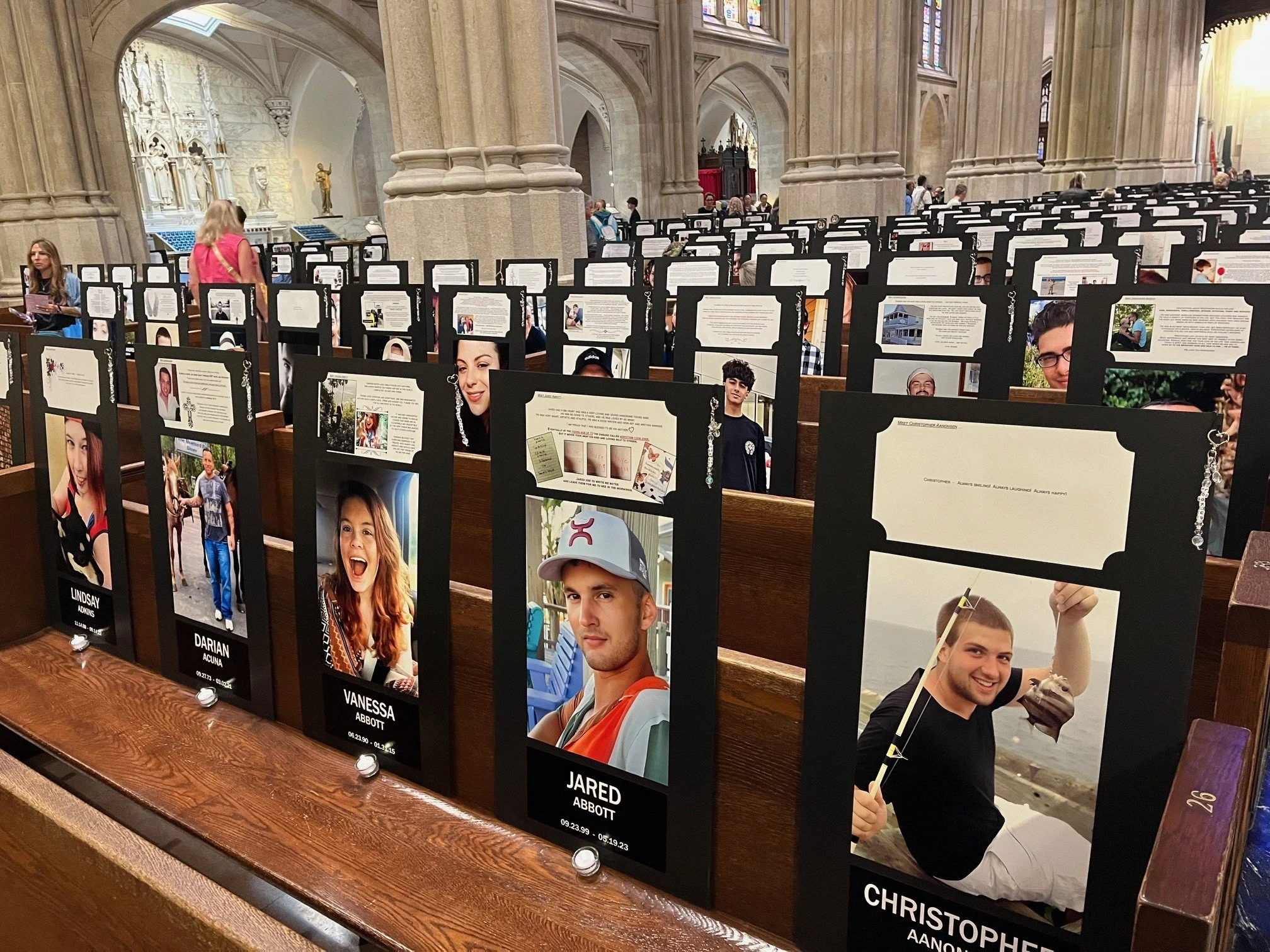 Indoor scene of a church with numerous memorial display boards featuring photographs and information about various individuals. The boards are placed on a wooden table, with some people viewing and reading them. The church's architecture includes lar