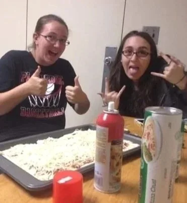 Two young women with glasses making silly faces and hand gestures next to a tray of shredded cheese, with bottles, a can, and a paper towel roll on a wooden table.
