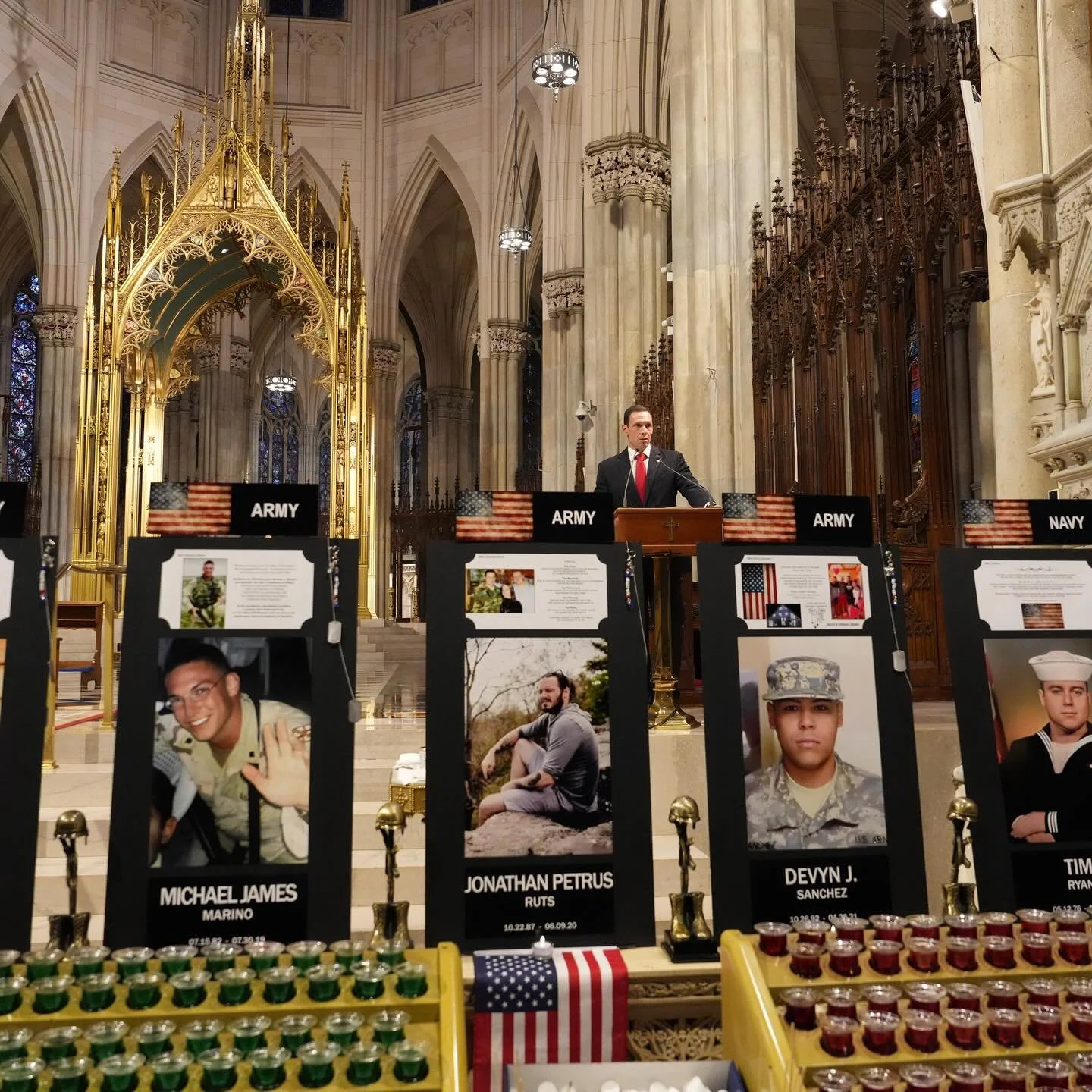 A man in a suit and red tie standing at a podium inside a grand, ornate church or cathedral with high arched ceilings and stained glass windows. In the foreground, there are display boards with photographs and names of military personnel, including M