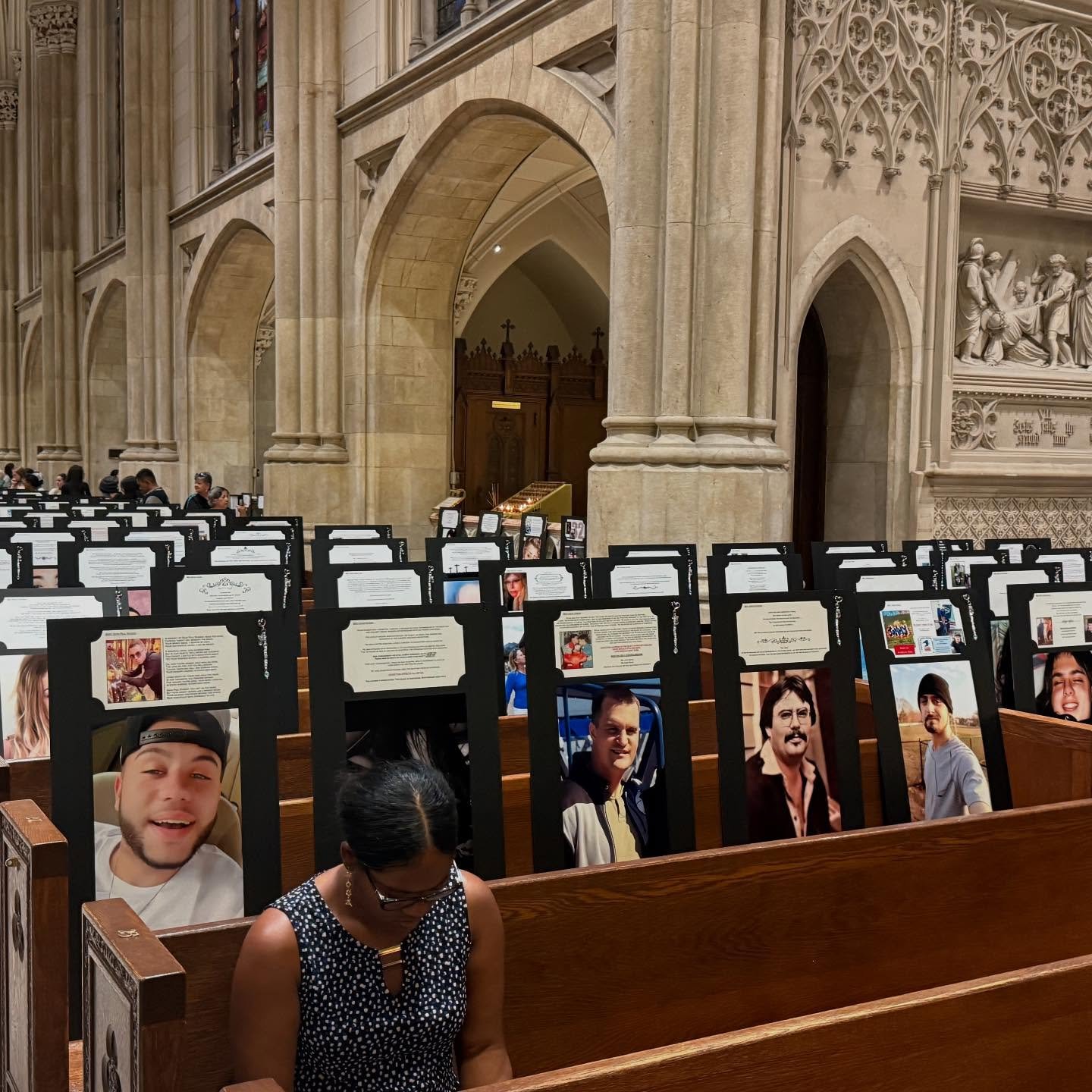 Inside a church with wooden pews decorated with photos and printed materials, some people sit, and there are large stone arches and detailed stone carvings in the background.