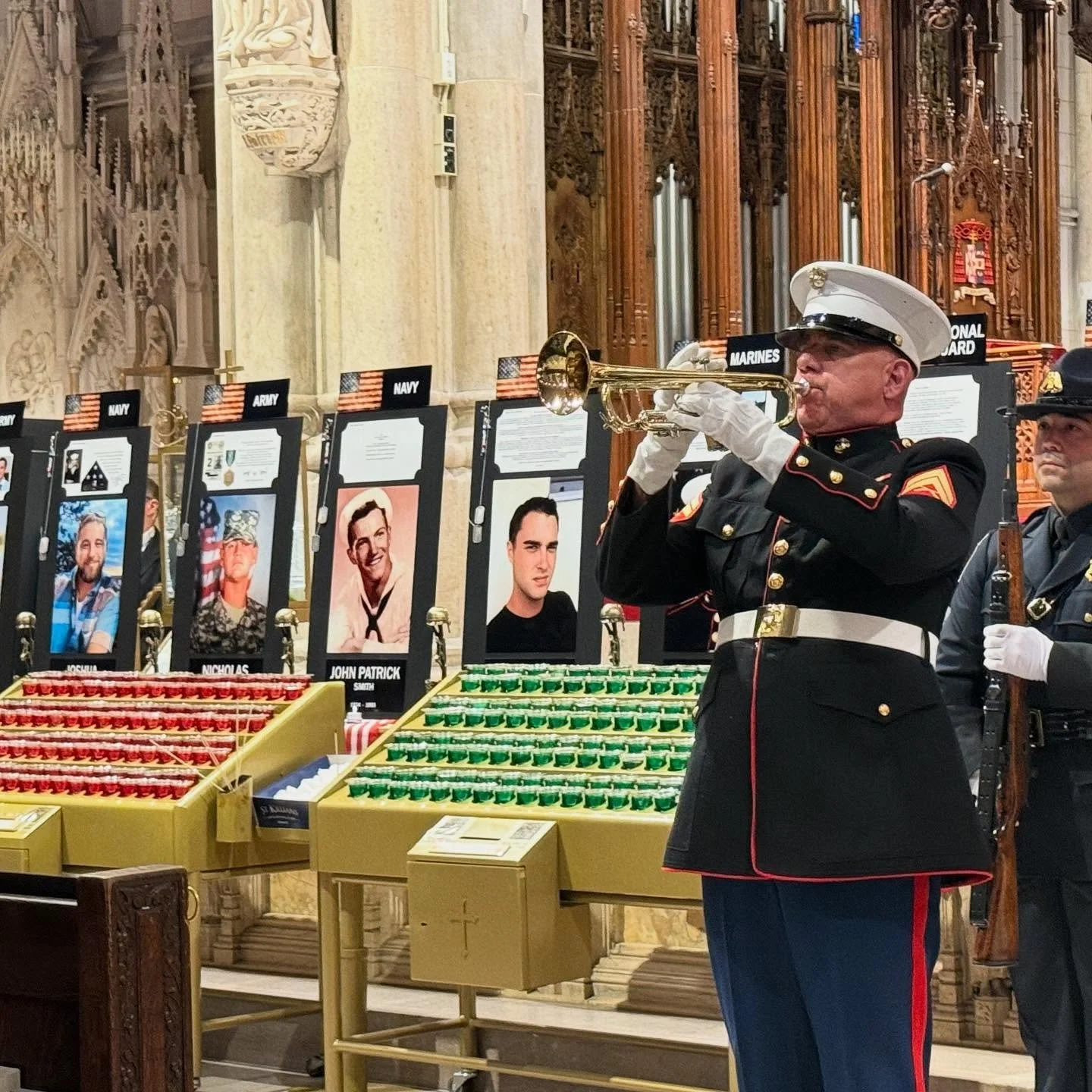 A military funeral or memorial service inside a church or cathedral features a Marine playing the bugle, dressed in full formal uniform with a white hat and white gloves. Behind him, another soldier holds a rifle. In the background, there are portrai