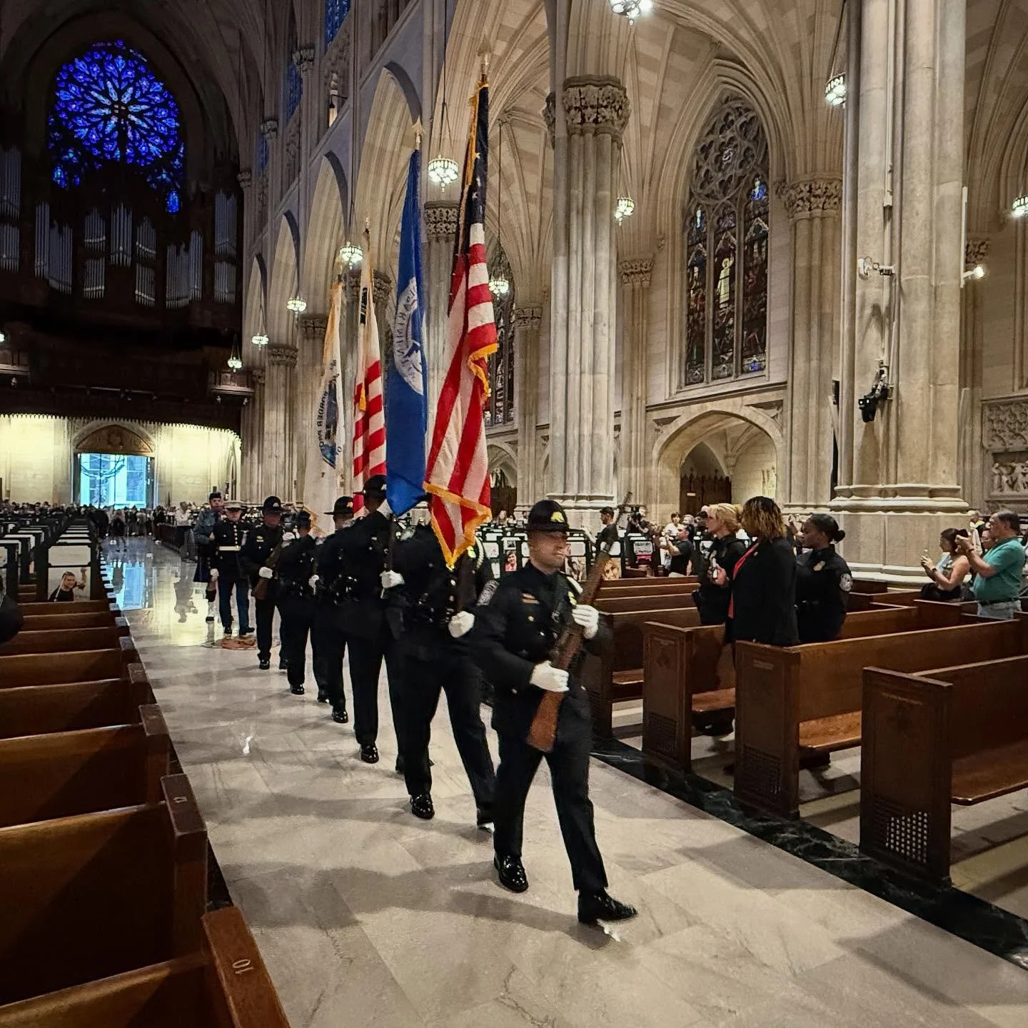 A procession of police officers carrying flags inside a large cathedral with stained glass windows and high vaulted ceilings.