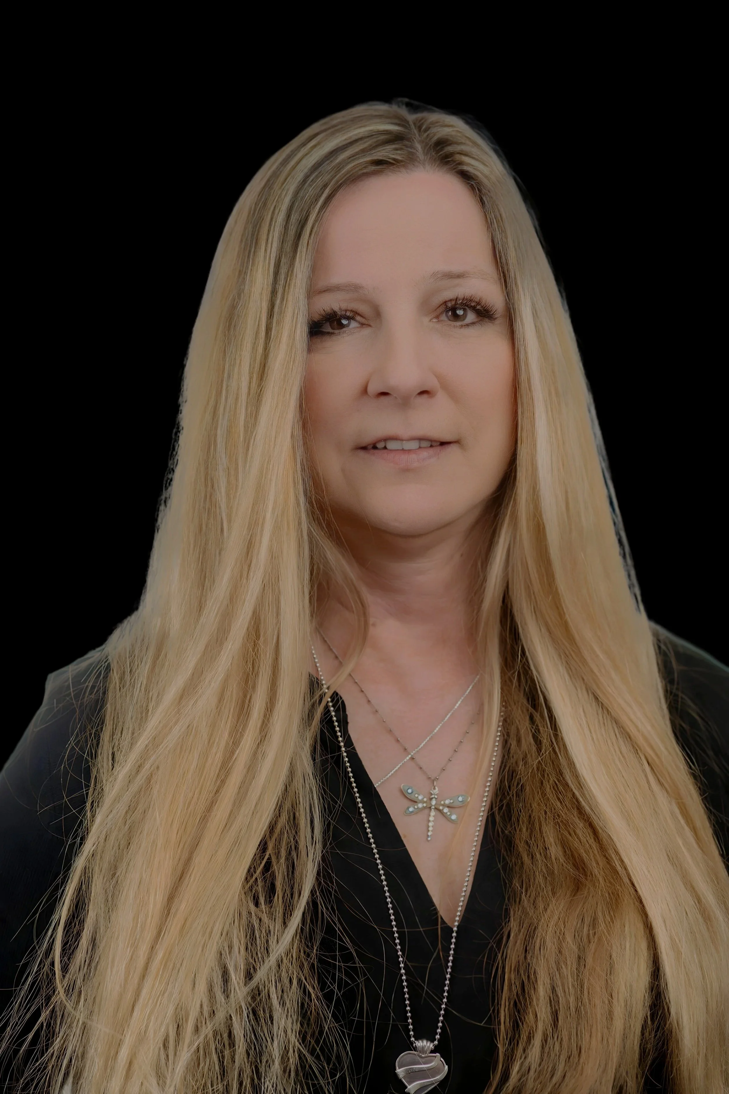 A woman with long blonde hair, light skin, wearing black top and layered necklaces including one with a dragonfly pendant, against a black background.