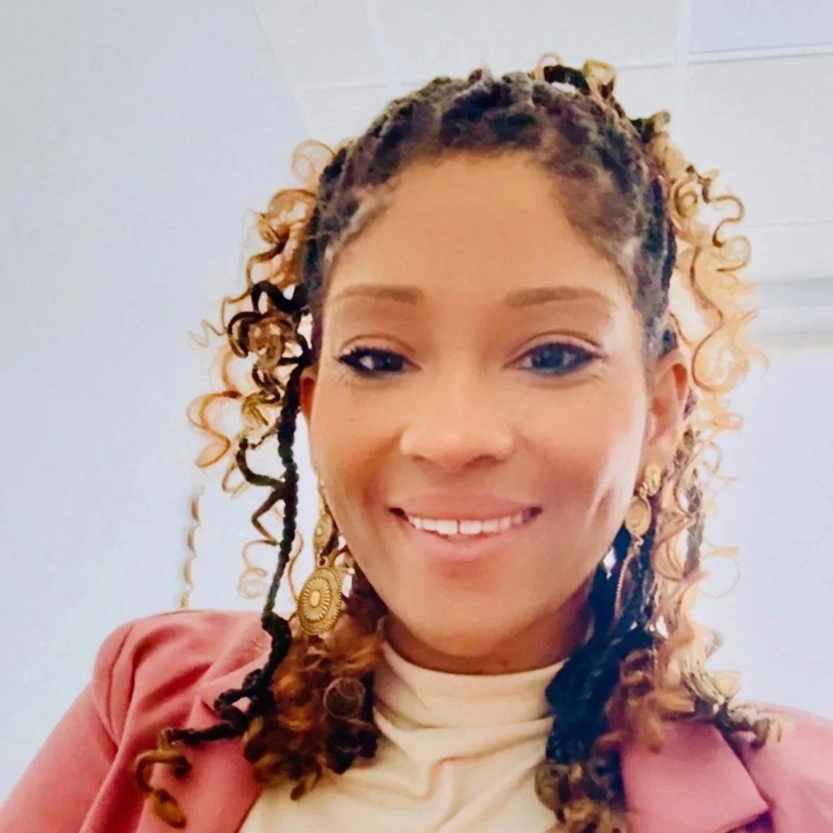 A woman with curly, blonde and brown styled hair, wearing earrings and a light-colored turtleneck, smiling at the camera.
