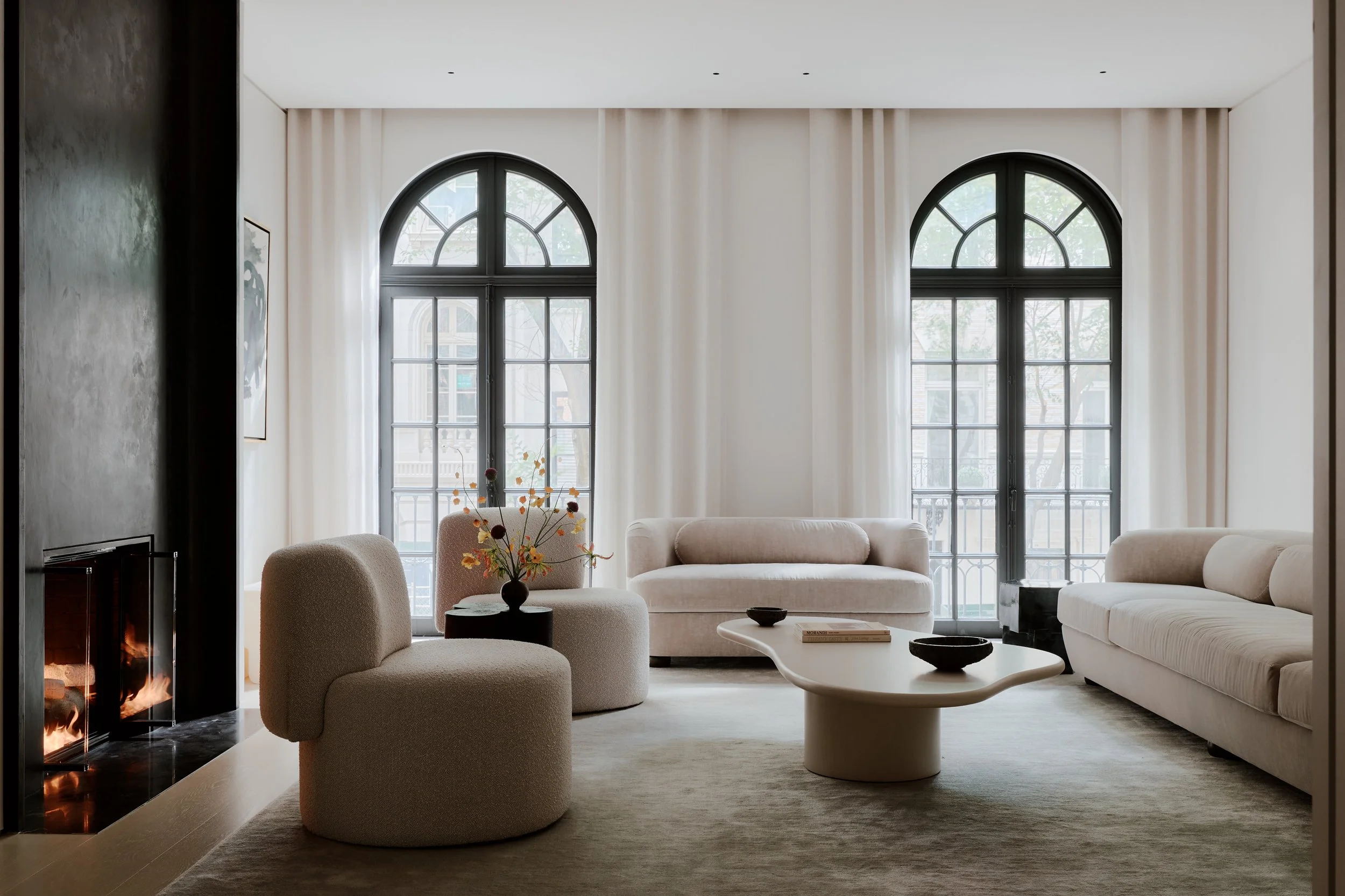 Modern living room with large arched windows, white sofas, a beige armchair, a black fireplace, and a coffee table with decorative bowls and a book, illuminated by natural light.