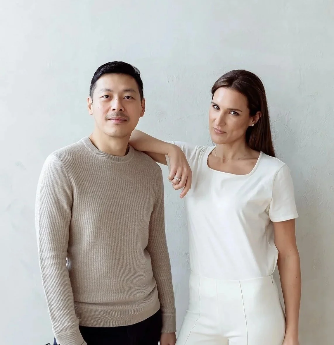 A man and a woman standing next to each other against a light-colored wall. The woman has her arm around the man's shoulder.