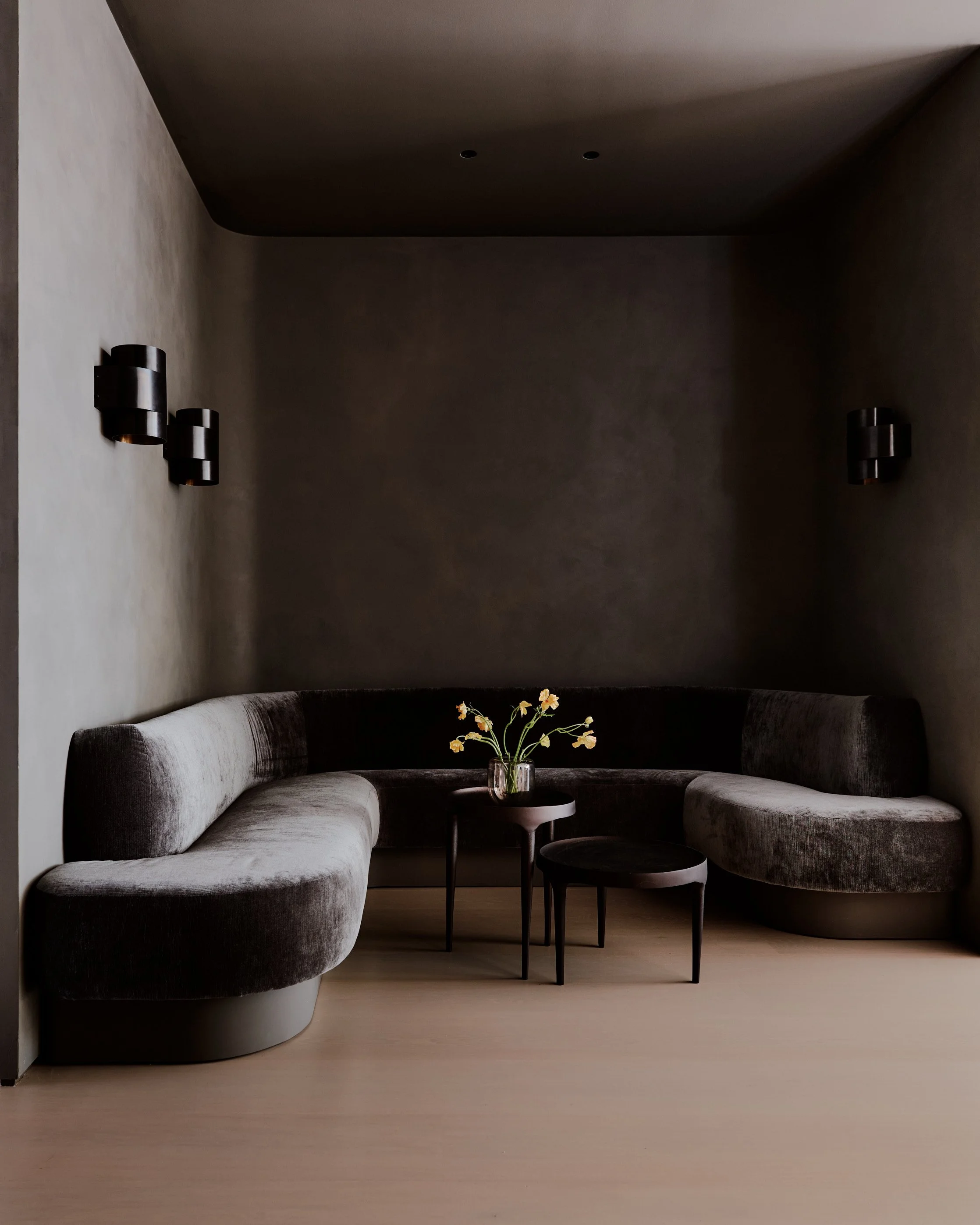 Living room corner with curved gray velvet sectional sofa, a small round black side table, a taller black round table, a clear glass vase with yellow flowers, a beige wood floor, dark brown walls, and black wall lamps.