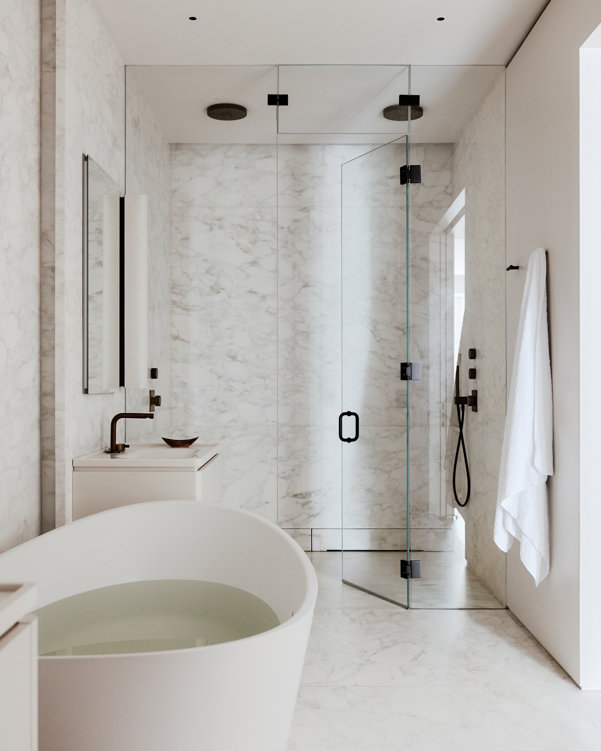 Modern bathroom with white marble walls and floor, glass shower enclosure, towel, and a white freestanding bathtub.