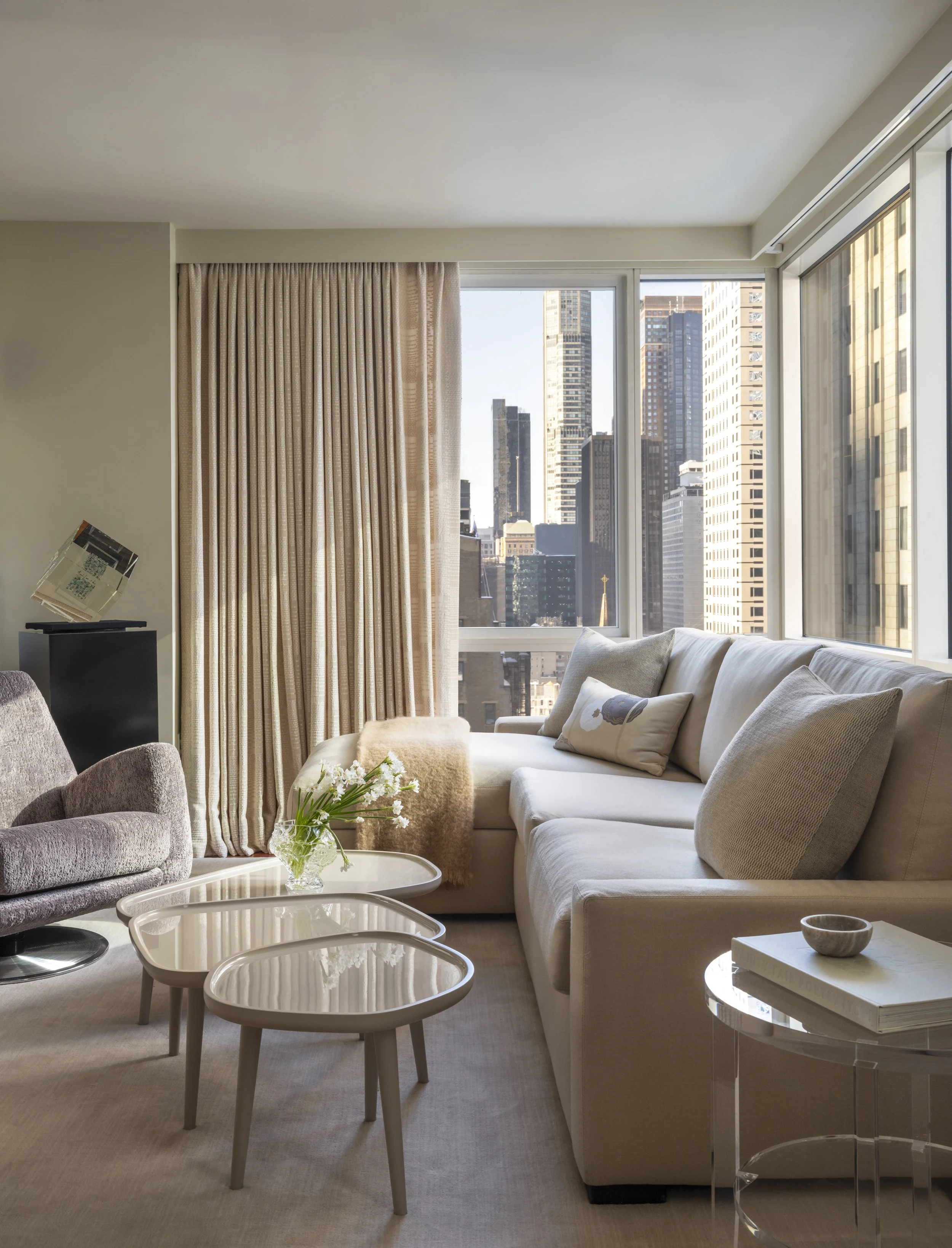 Living room with beige and white furniture, large windows showing city skyscrapers, beige curtains, glass coffee tables, and decorative items.