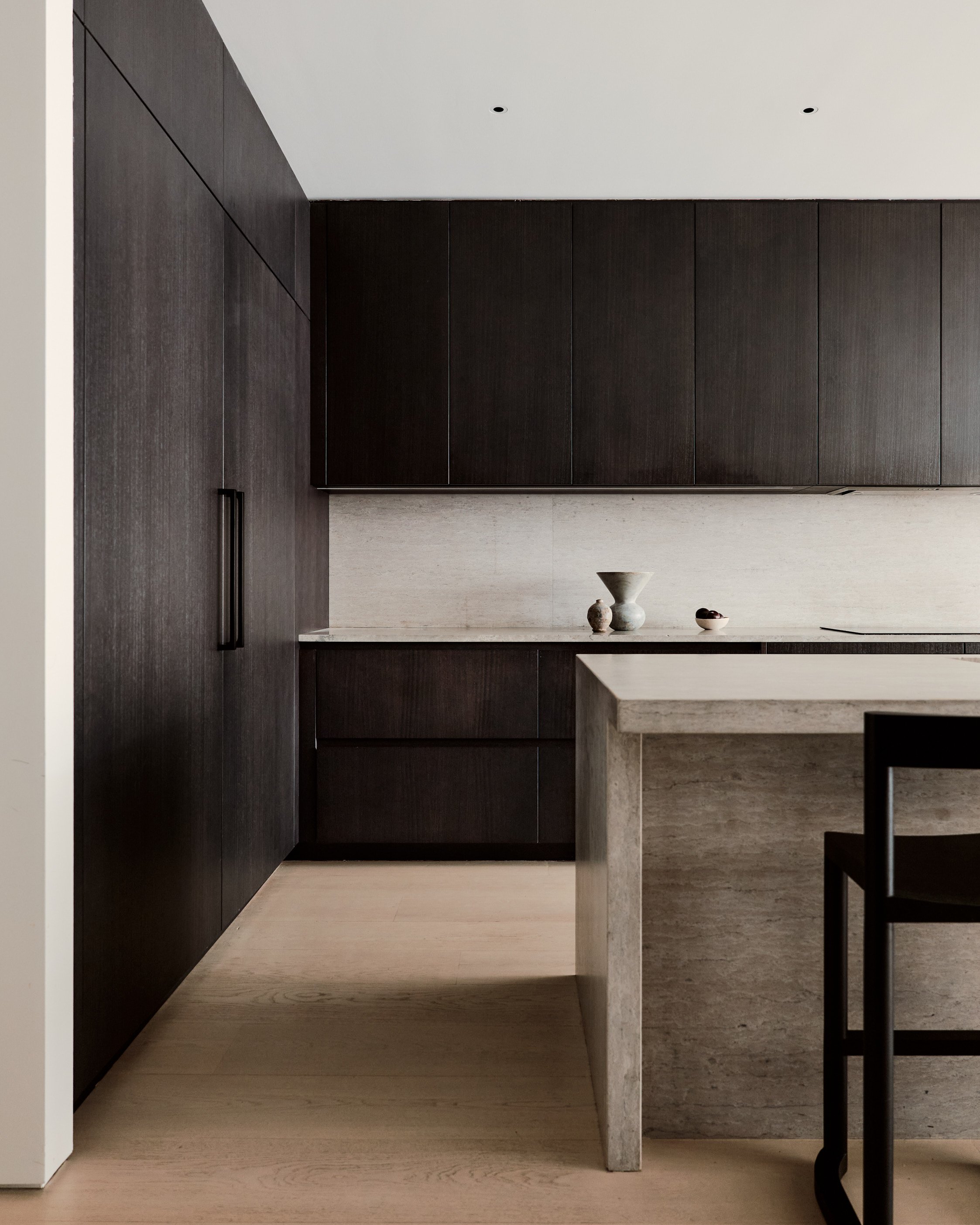 A modern kitchen with dark wooden Italian cabinets, a beige marble island, and decorative vases on the counter.