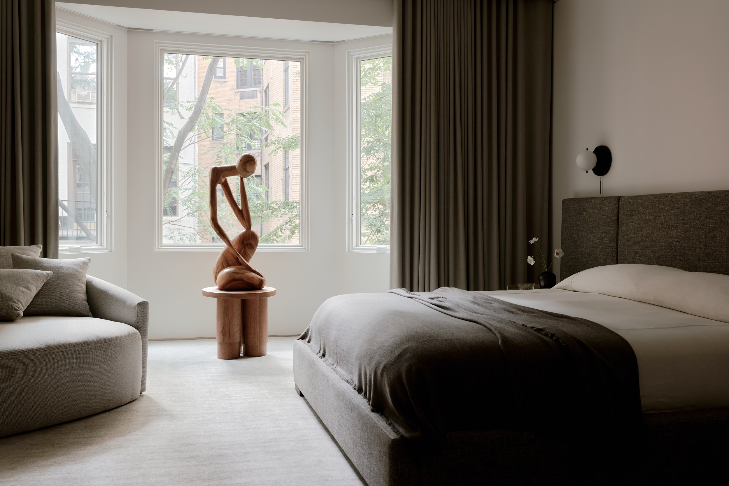 A modern bedroom with large windows, a grey upholstered bed, a side table with a wooden sculpture, and a small flower vase. Dark curtains frame the windows, and outside trees are visible.