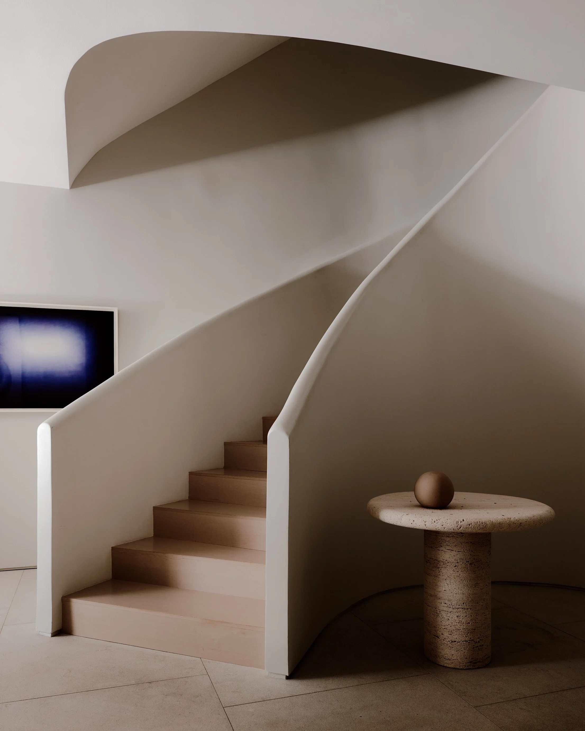 Empty modern staircase with white curved railings leading up to a small landings, adjacent to a round pedestal table with a beige stone sphere on it, in a minimalist room with light-colored walls and flooring.