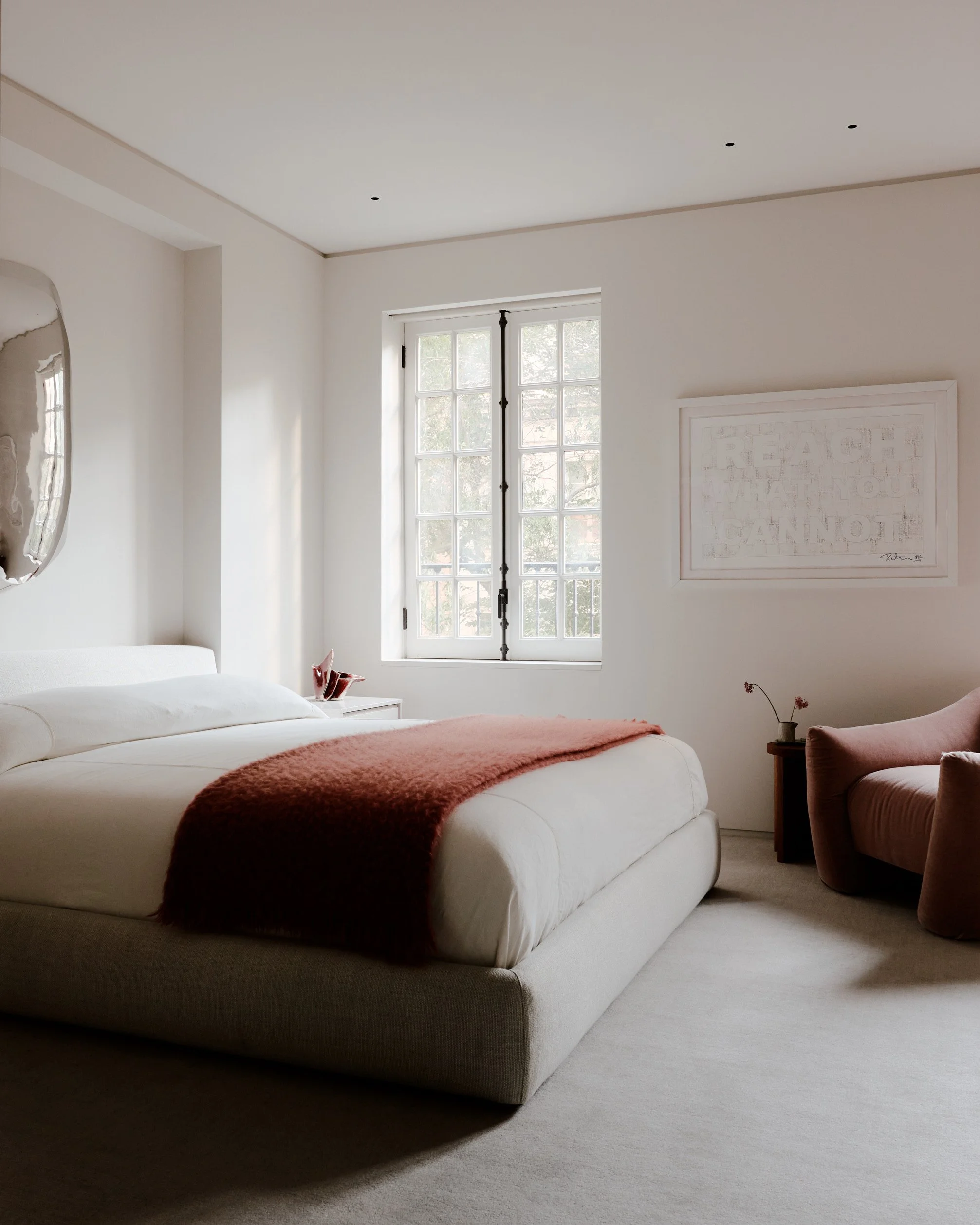 A bedroom with a large bed covered with a white sheet and a red blanket, a small side table with a decorative item, a pink armchair, and a window with glass panes letting in natural light.