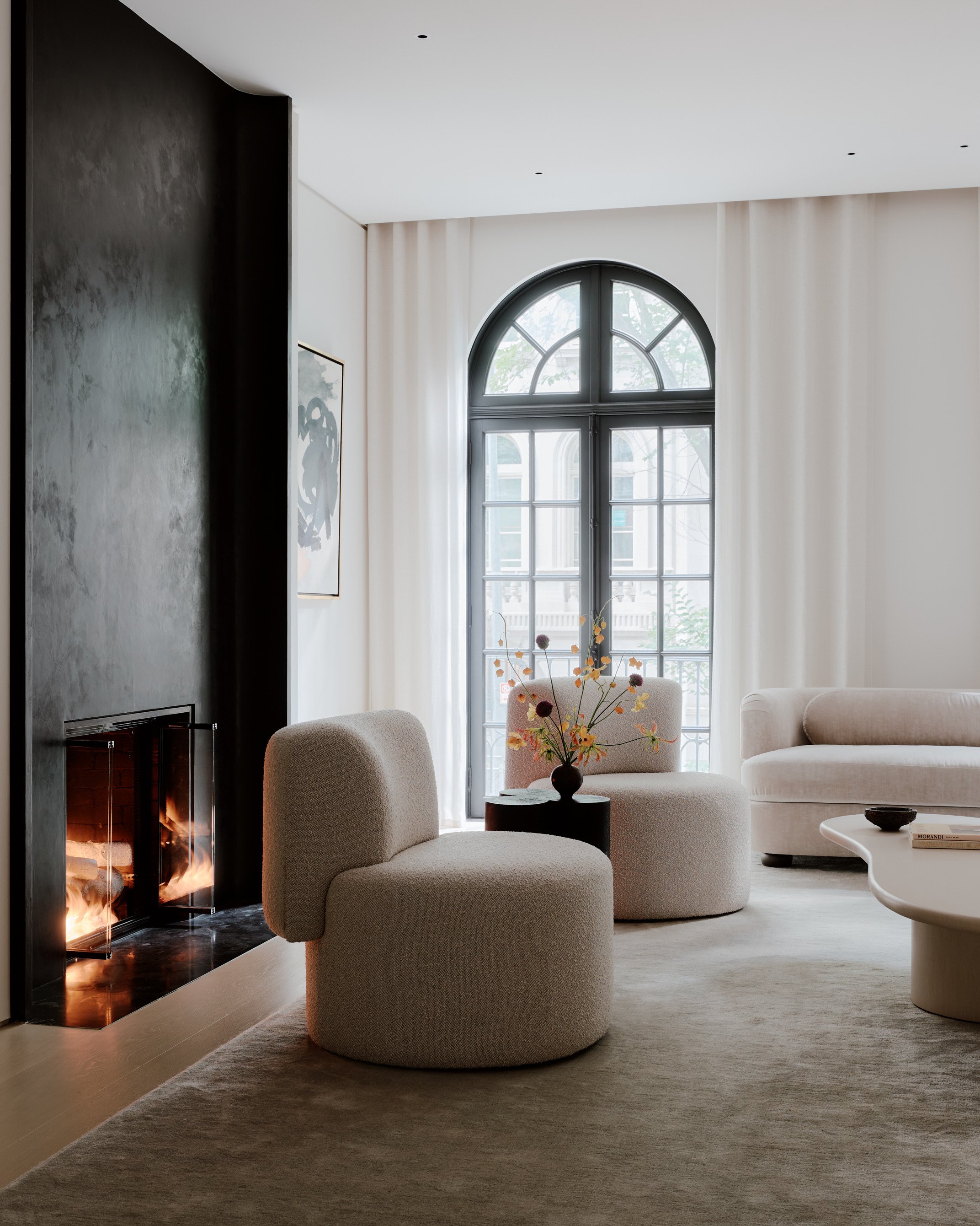 A cozy living room with a modern fireplace, two beige armchairs, a white sofa, a round coffee table, and a large arched window with curtains. A vase of flowers sits on a side table, and a book is on the coffee table.