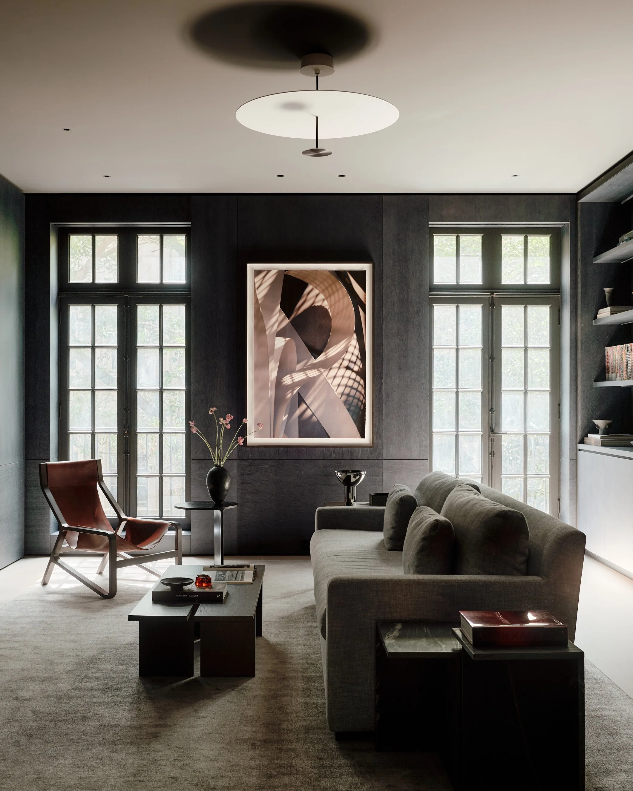 A modern living room with large windows, a gray couch, a black coffee table with books and decorative items, a chair, a unique ceiling light fixture, artwork on the wall, and various decorative objects.